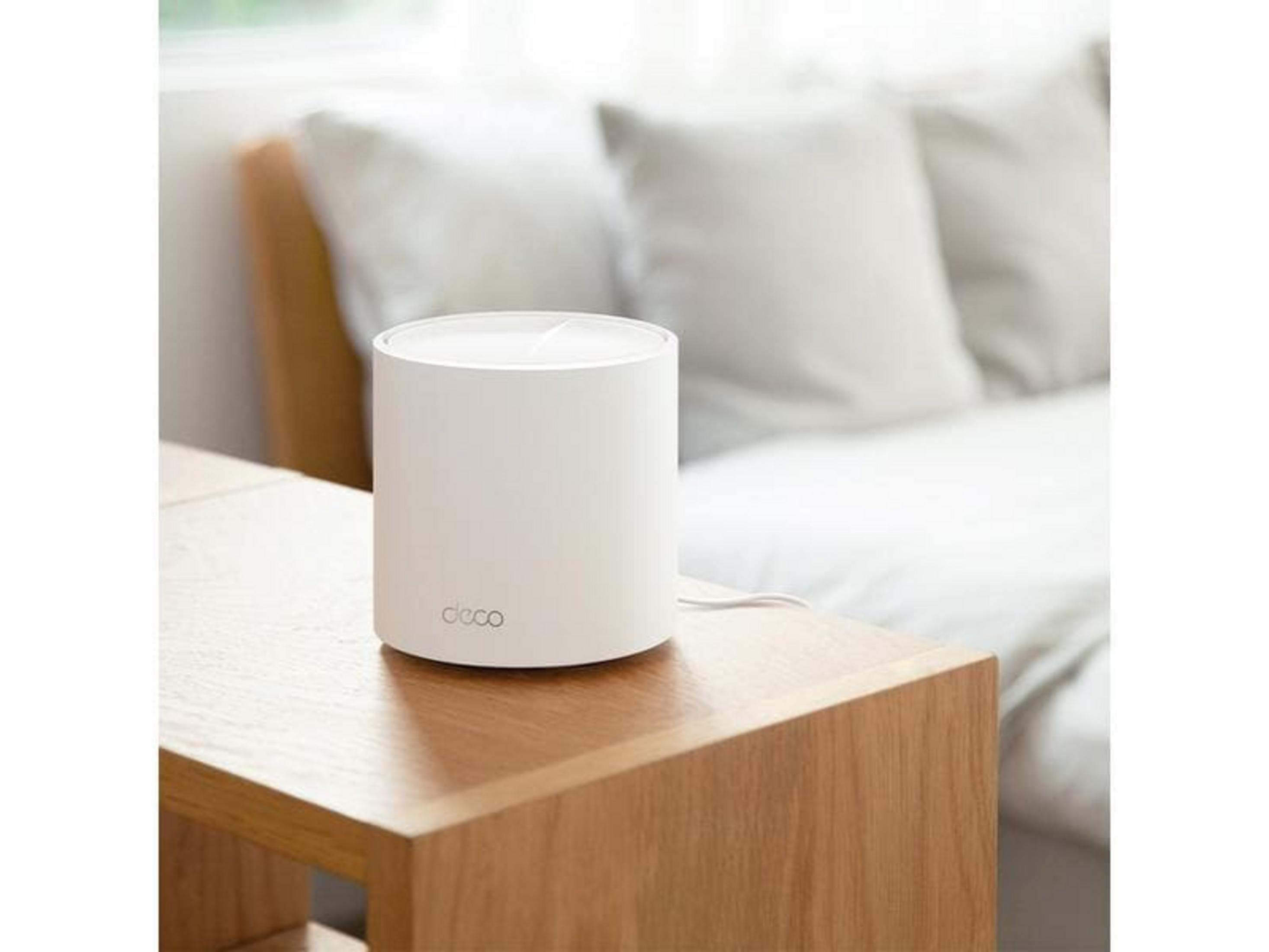 Biały, cylindryczny router stoi na drewnianym stole przed łóżkiem z białymi poduszkami.