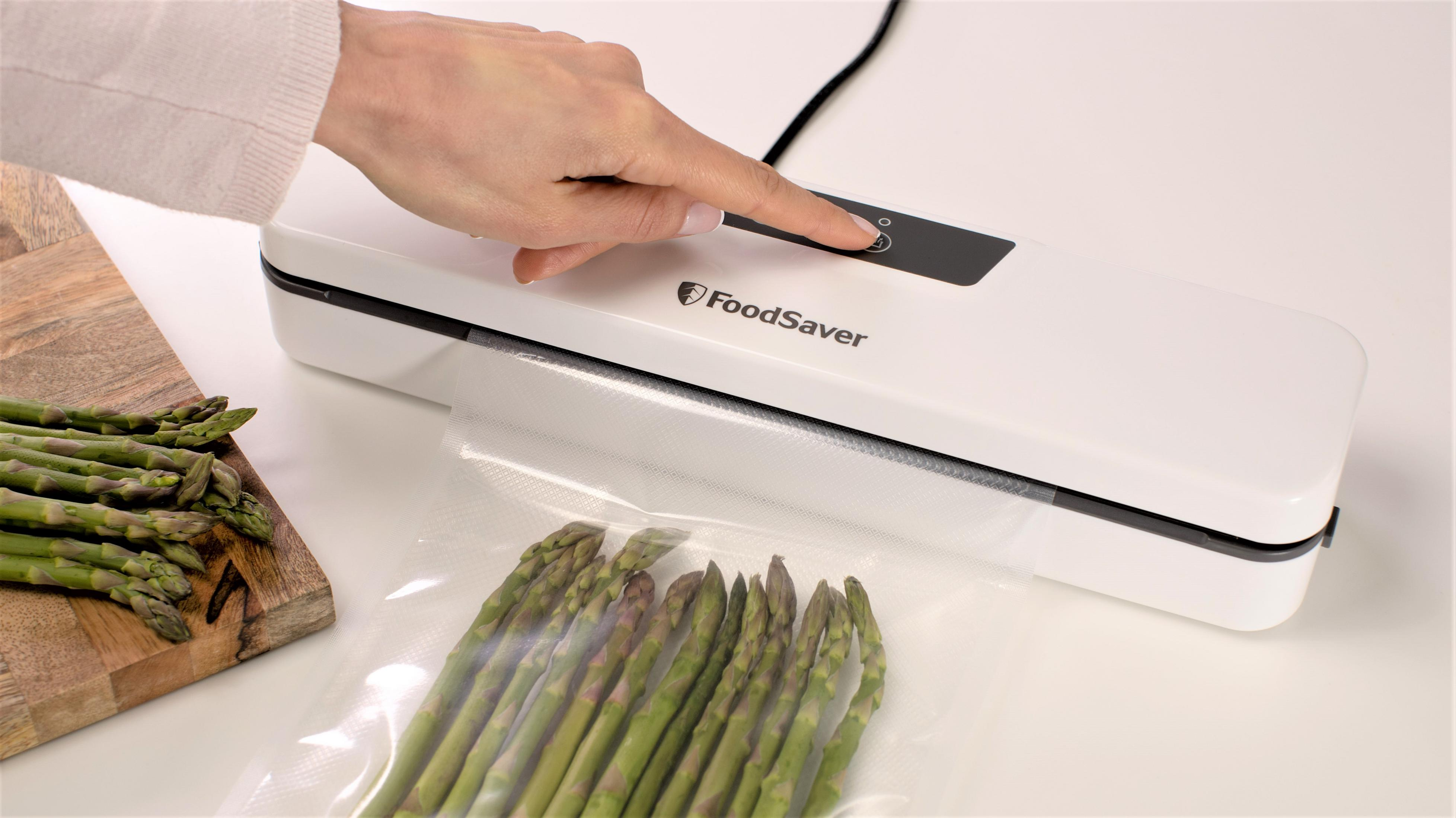 Ręka naciska przycisk na białej zgrzewarce próżniowej obok zielonych szparagów.