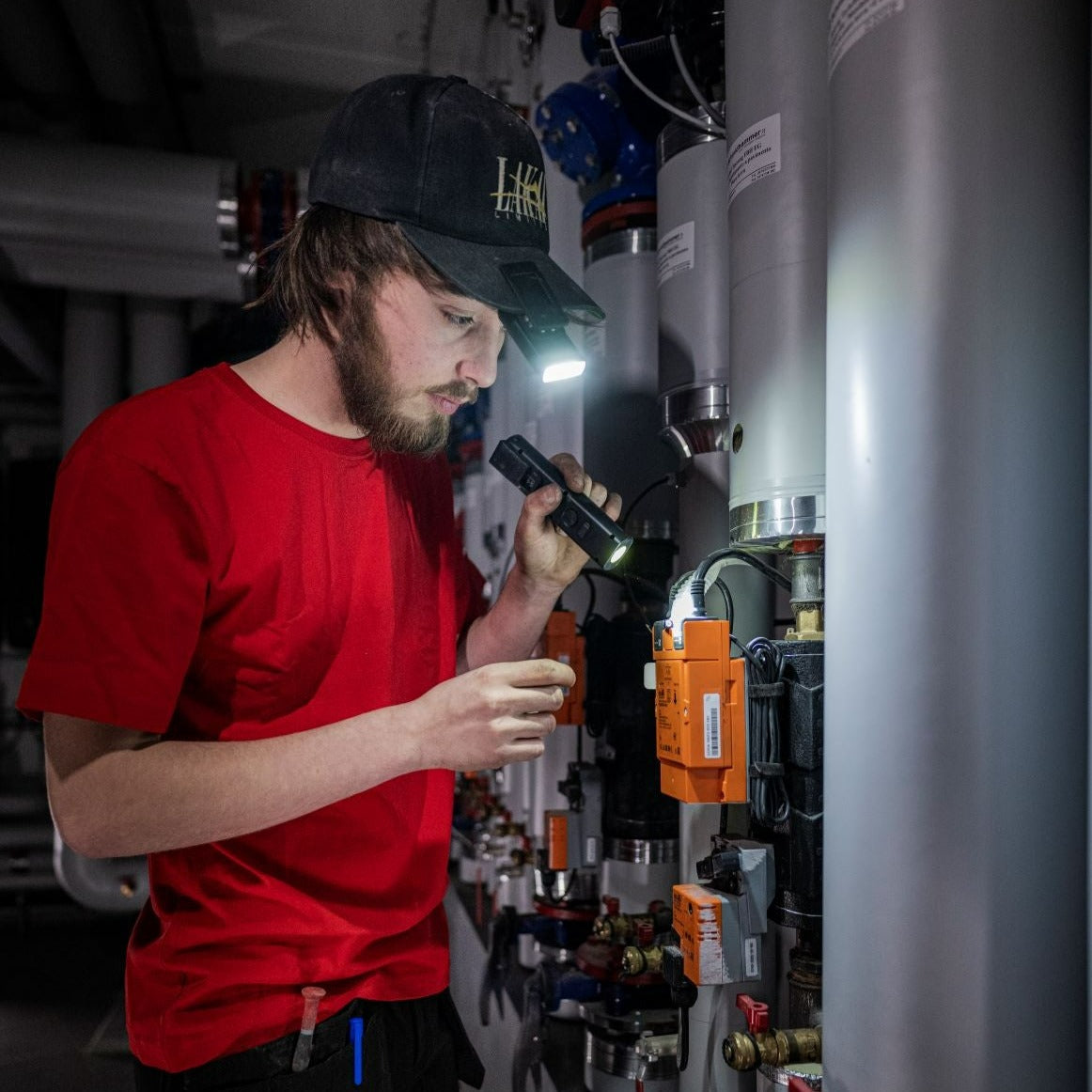 Un homme en chemise rouge et casquette inspecte des tuyaux avec une lampe.