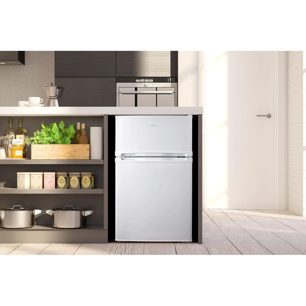 Ein weißer Kühlschrank steht in einer Küche mit Regalen voller Lebensmittel. Im Hintergrund ist eine Tür.