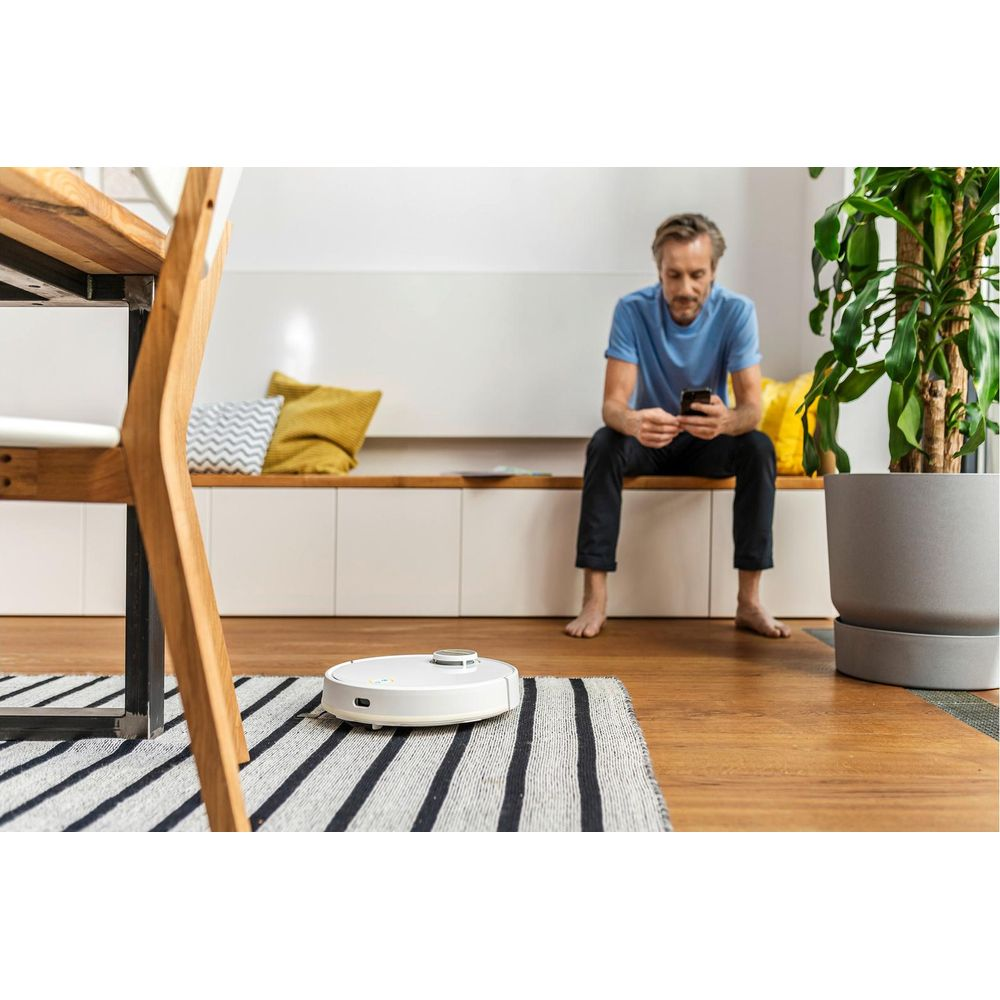 Biały robot odkurzający czyści dywan, podczas gdy mężczyzna siedzi na ławce.