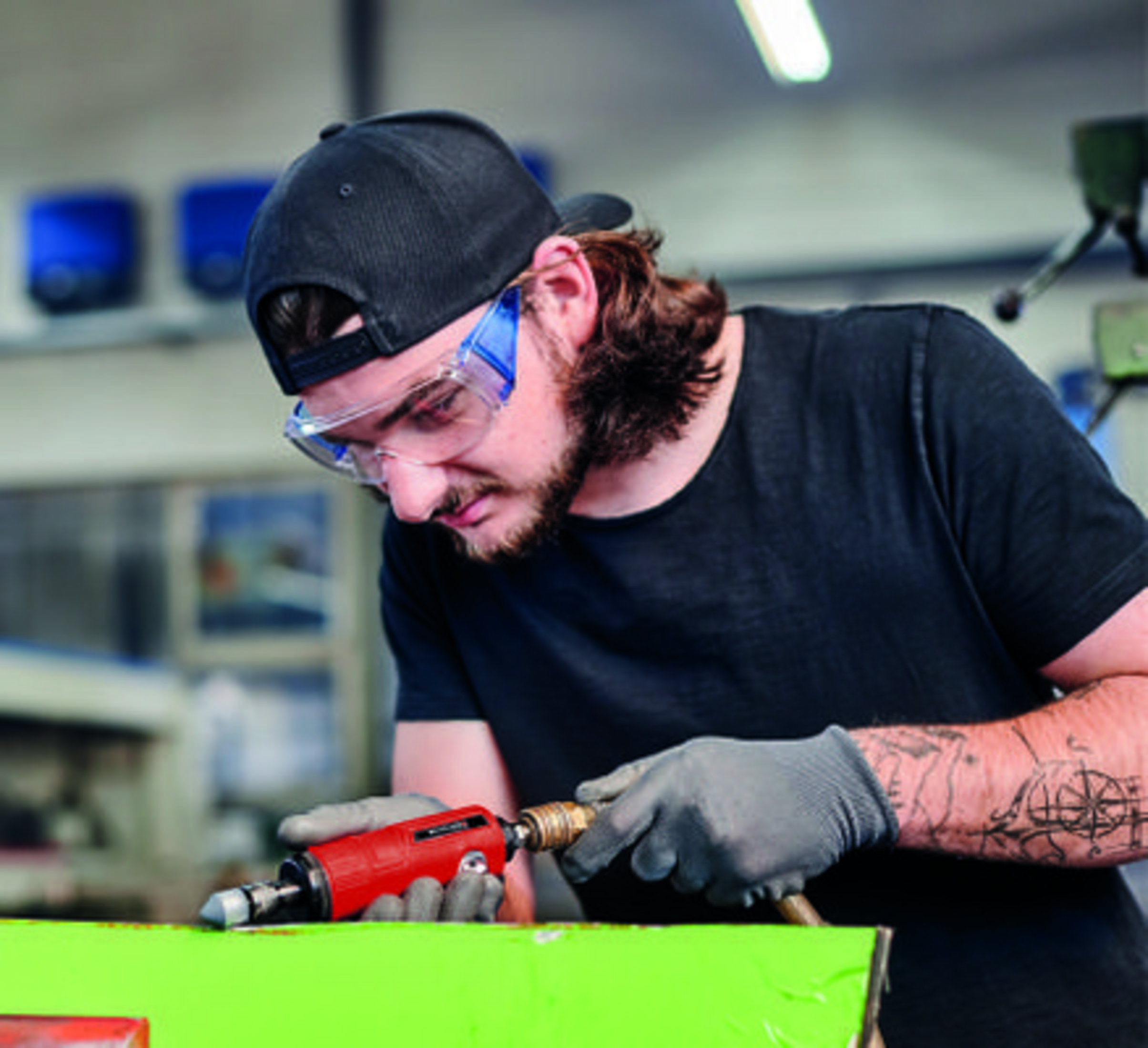 Man met veiligheidsbril werkt aan groen object met gereedschap.
