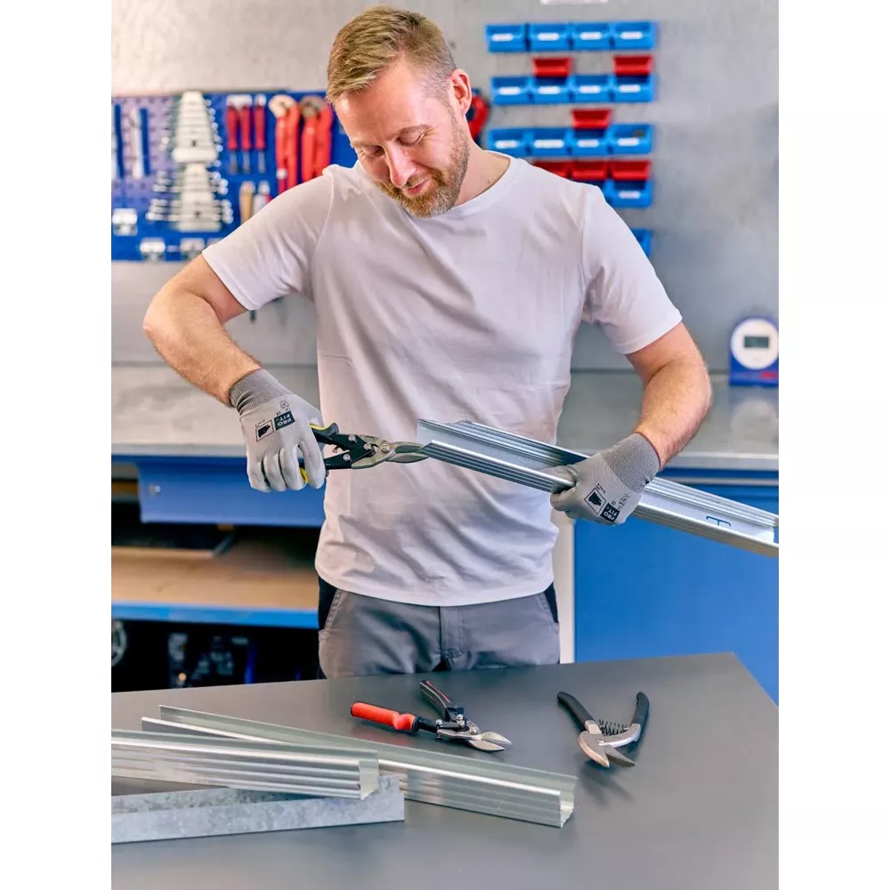 Mann mit Handschuhen schneidet Metallprofil mit Schere, Werkzeuge liegen auf Tisch.