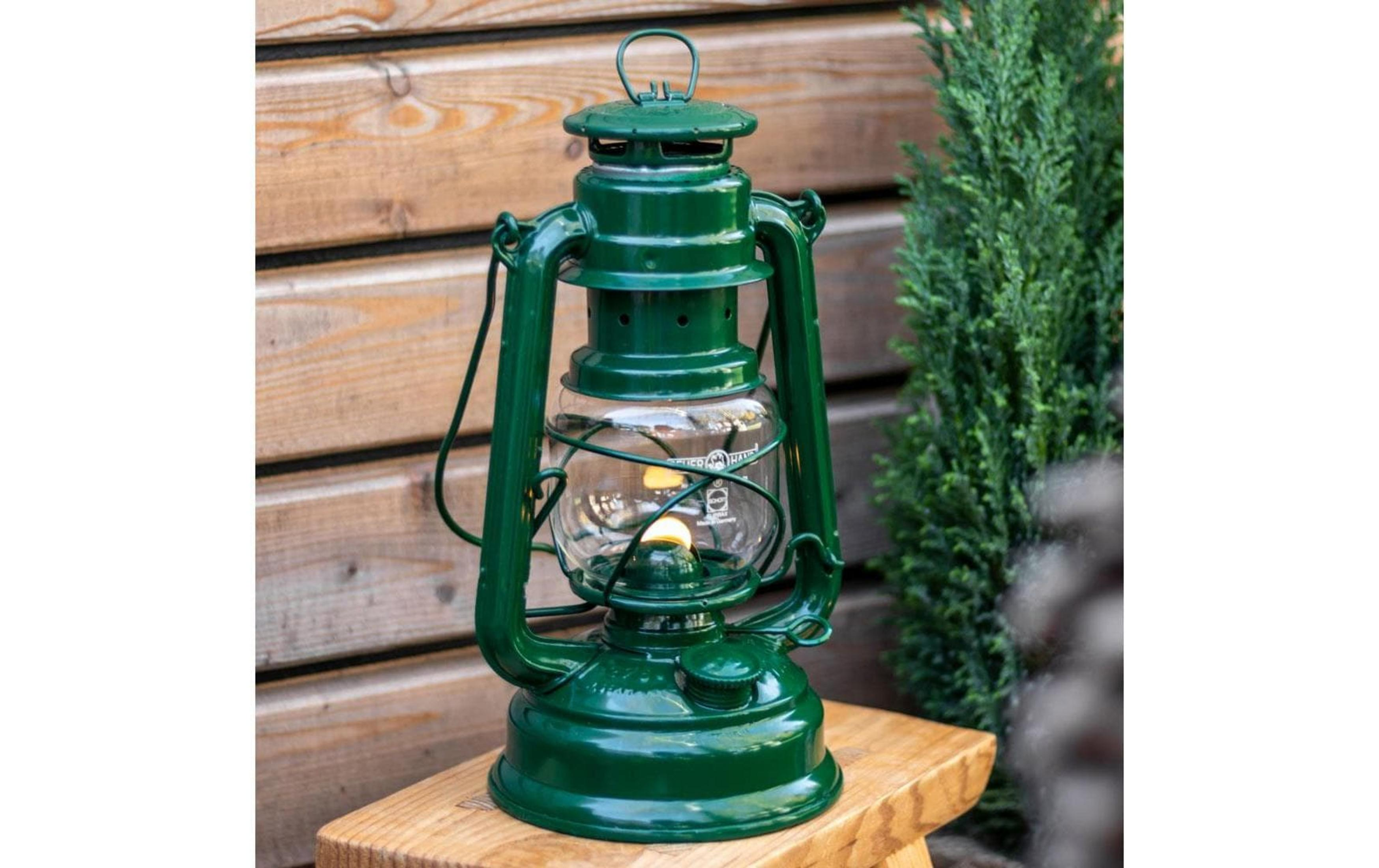 Eine grüne Petroleumlampe steht auf einem Holztisch vor einer Holzwand und einer Zypresse.