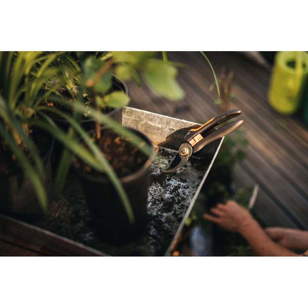 Gartenschere liegt auf einem Tisch neben Topfpflanzen. Hände sind im Hintergrund zu sehen.