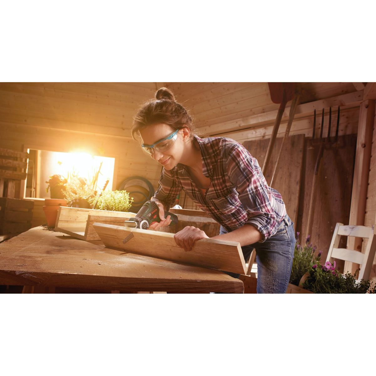 Kobieta w warsztacie piłuje drewno, nosi okulary ochronne i koszulę w kratę.
