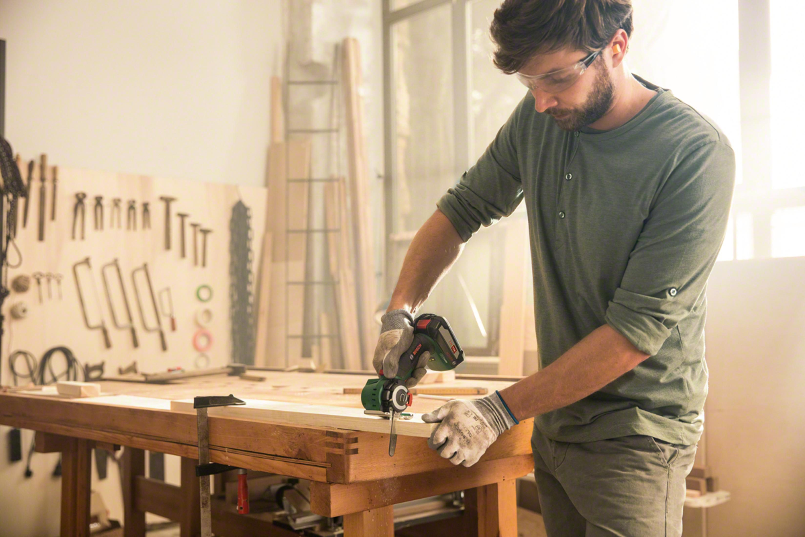 Mężczyzna w okularach i rękawicach piłuje drewno na ławce.
