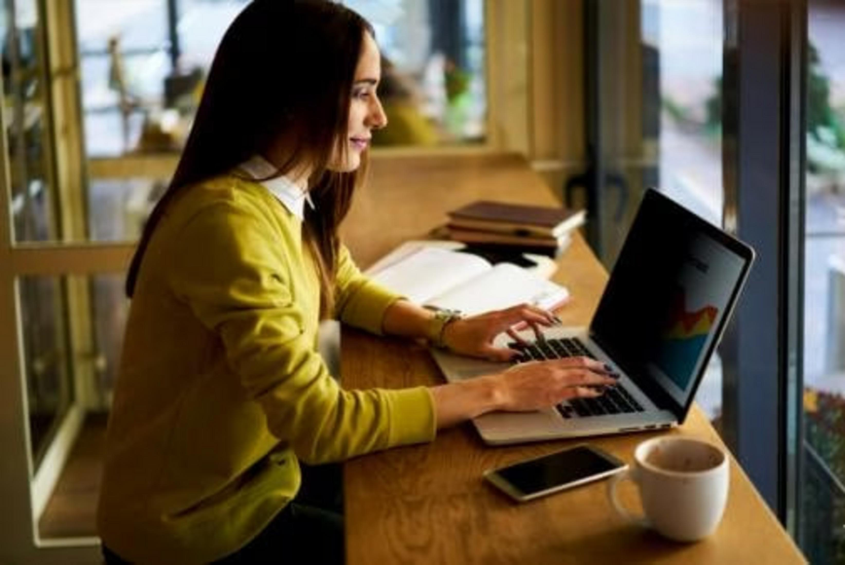 Eine Frau tippt auf einem Laptop in einem Café, neben ihr ein Handy und eine Tasse Kaffee.