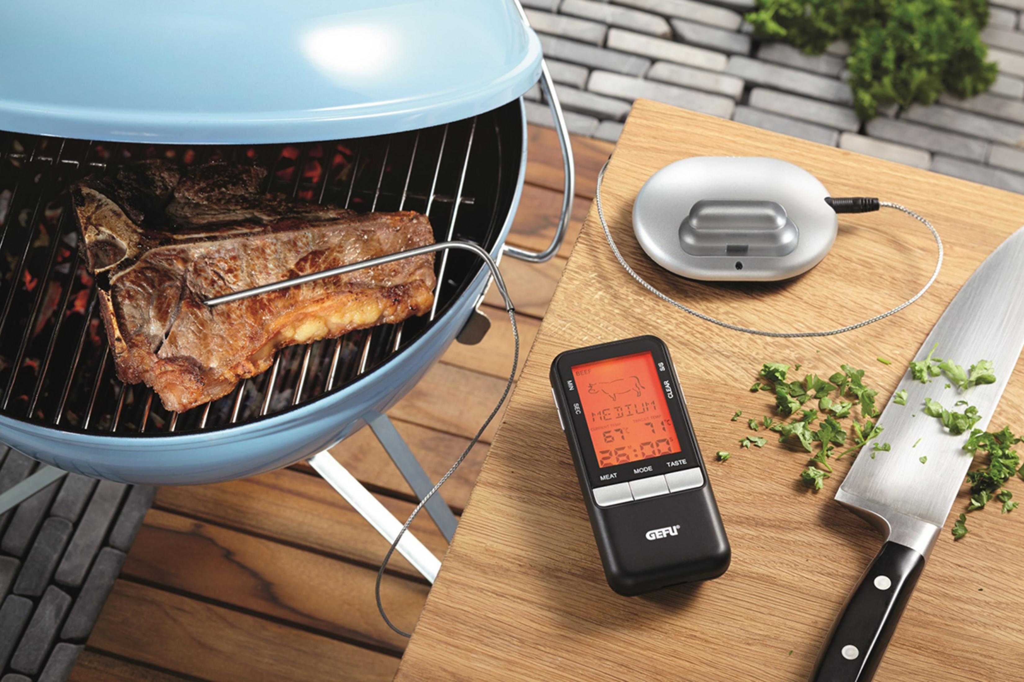 Ein T-Bone-Steak wird auf einem Grill zubereitet. Daneben liegen ein Fleischthermometer und ein Messer.