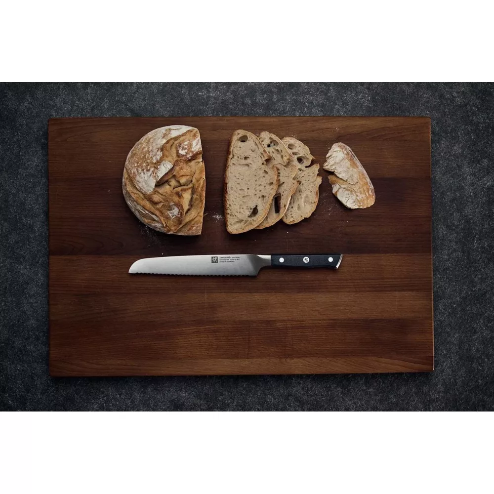 Nóż z ząbkowanym ostrzem leży na drewnianej desce obok bochenka i kromek chleba.