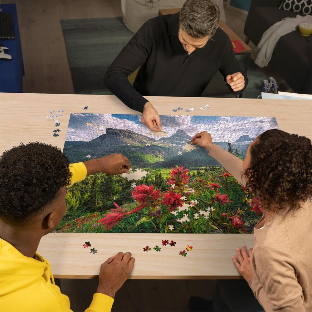 Drei Personen puzzeln ein Berglandschaftsbild mit roten Blumen im Vordergrund.