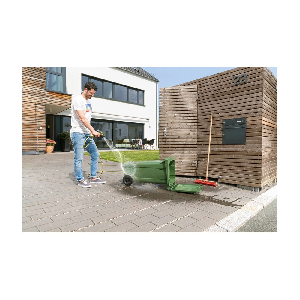 Mann reinigt grüne Mülltonne mit Gartenschlauch vor Holzhaus.