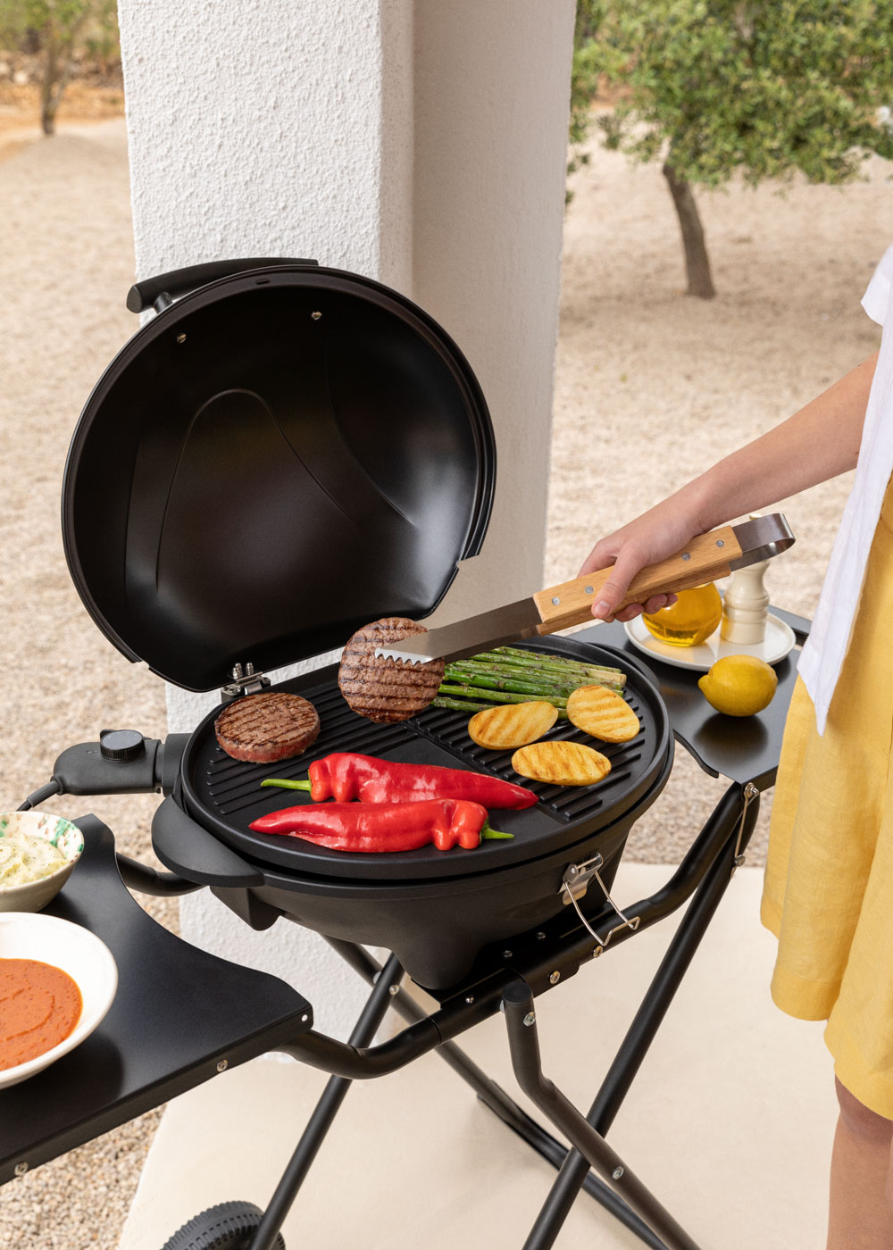 Una persona asa hamburguesas, espárragos, patatas y pimientos en una parrilla negra.