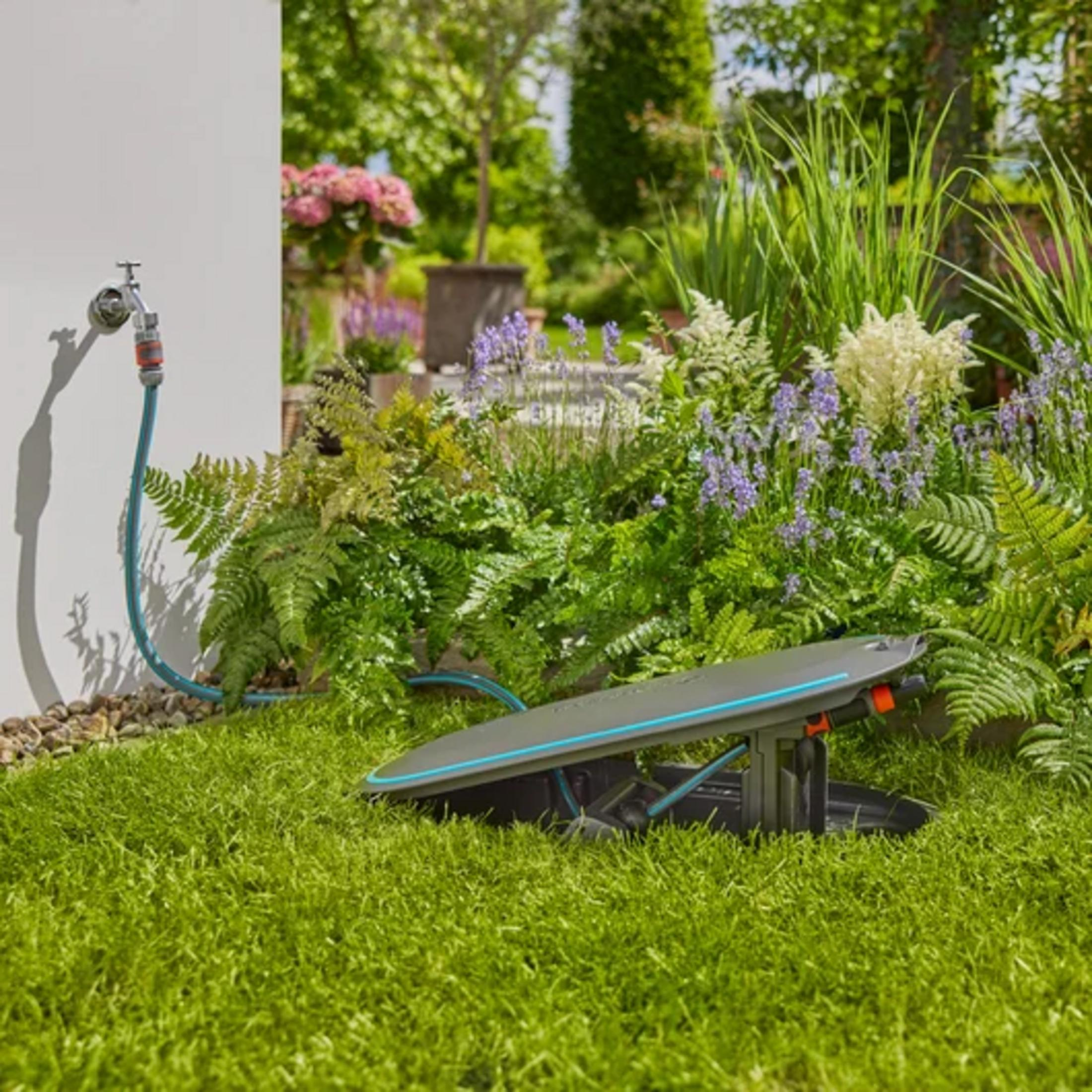 Un irrigatore da prato si trova su erba verde accanto a un giardino fiorito.