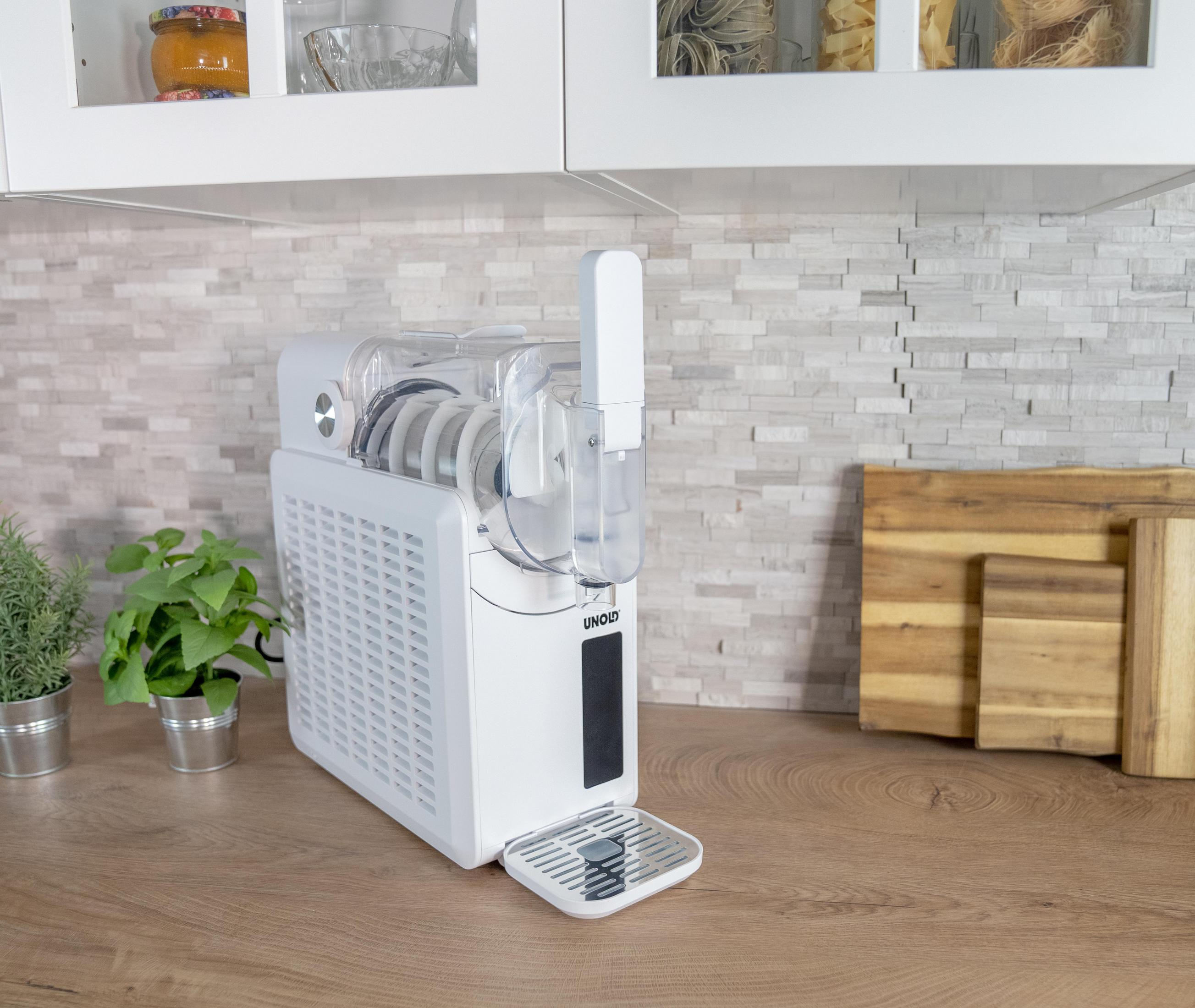 Biała maszynka do lodów stoi na drewnianej powierzchni obok dwóch doniczek z ziołami.