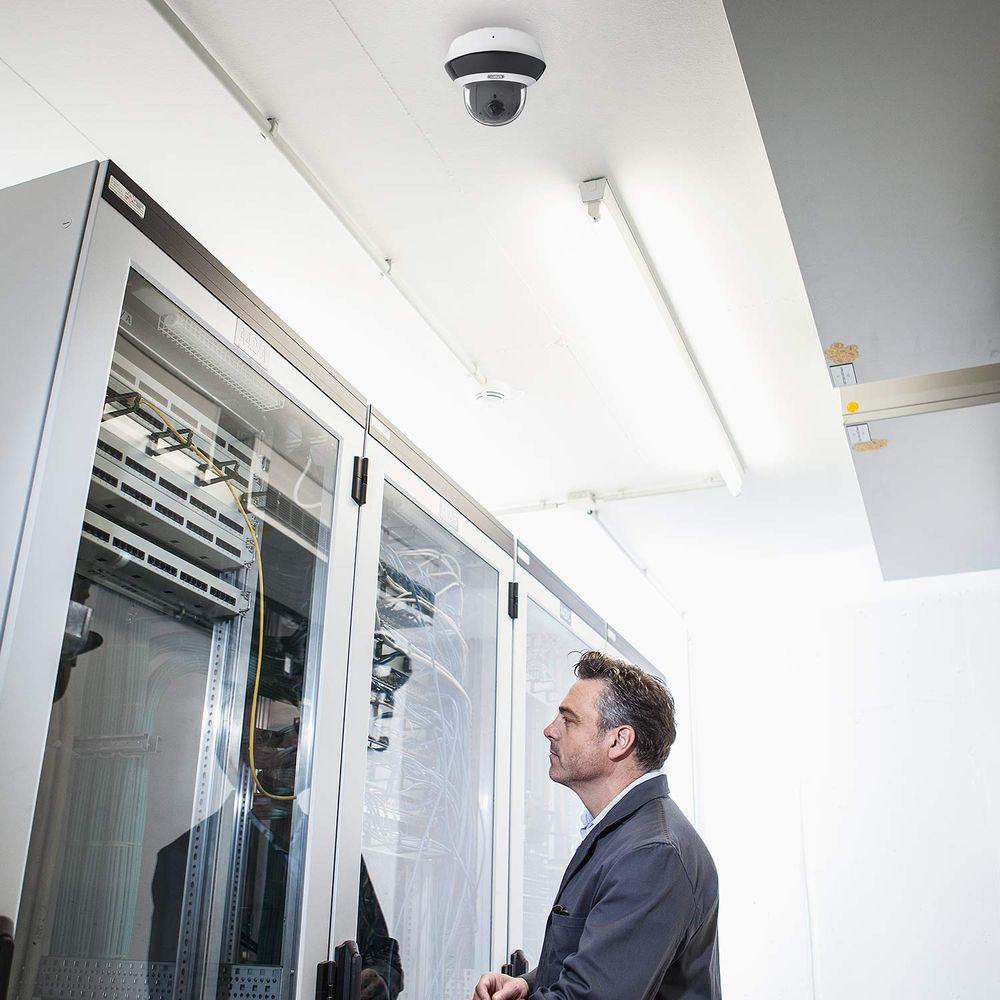 Mann blickt auf Server-Racks; Überwachungskamera an der Decke.