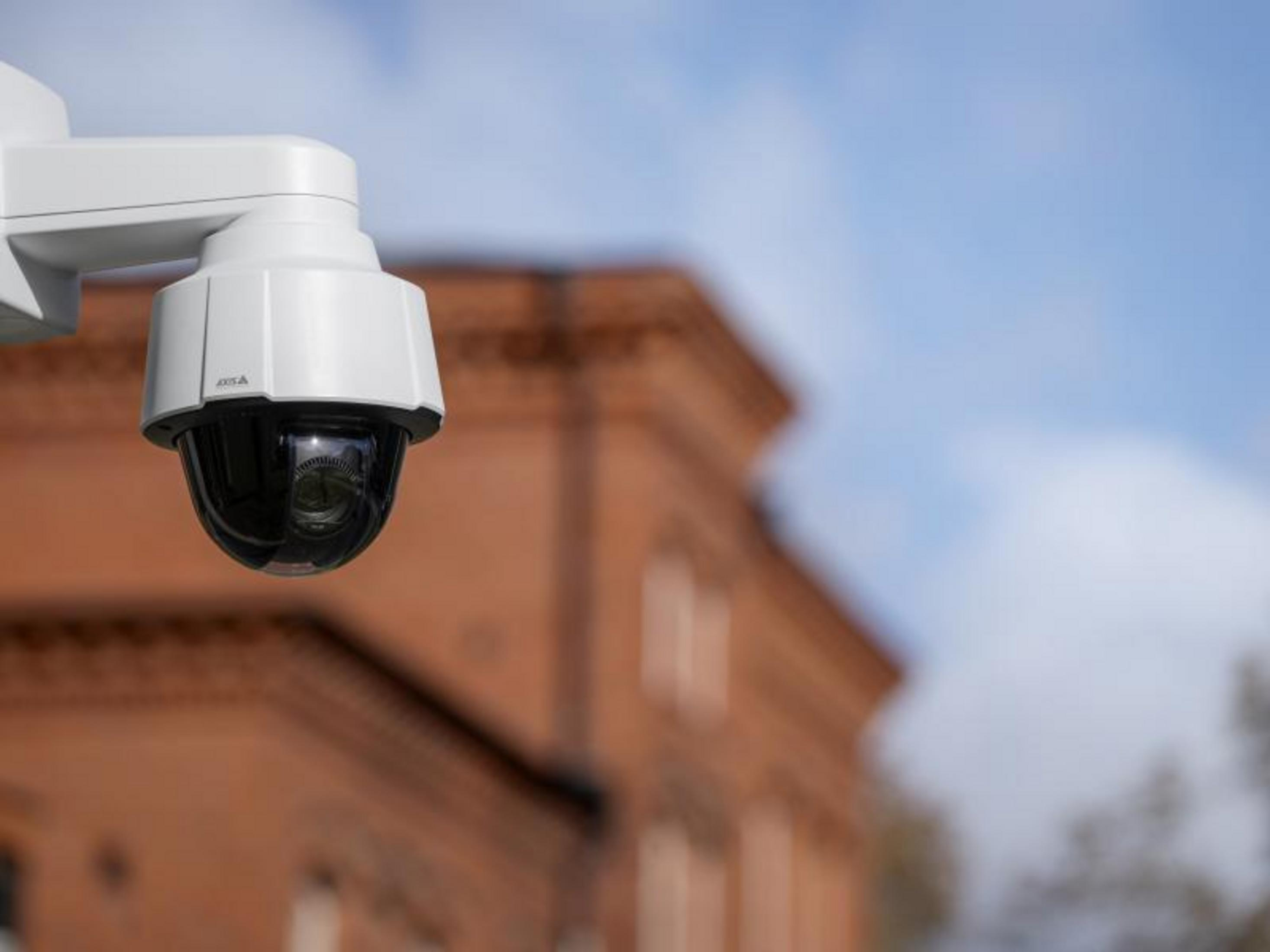 Biała kamera monitorująca z czarnym obiektywem przed ceglanym budynkiem.