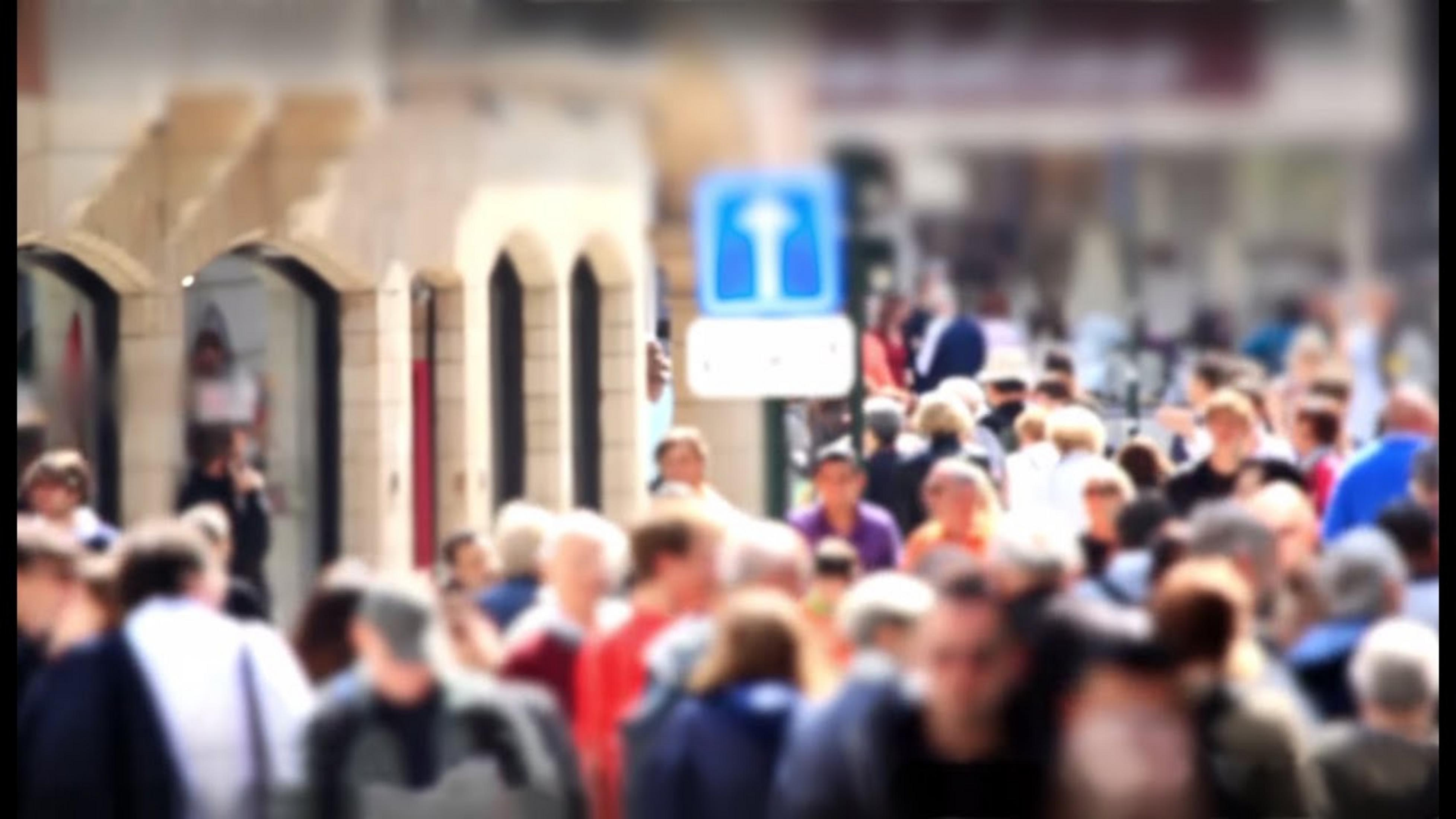 Tłum ludzi na ruchliwej ulicy z budynkami i niebieskim znakiem.