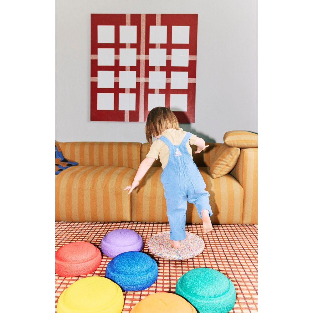 Un enfant en salopette bleue s'équilibre sur des coussins colorés devant un canapé.