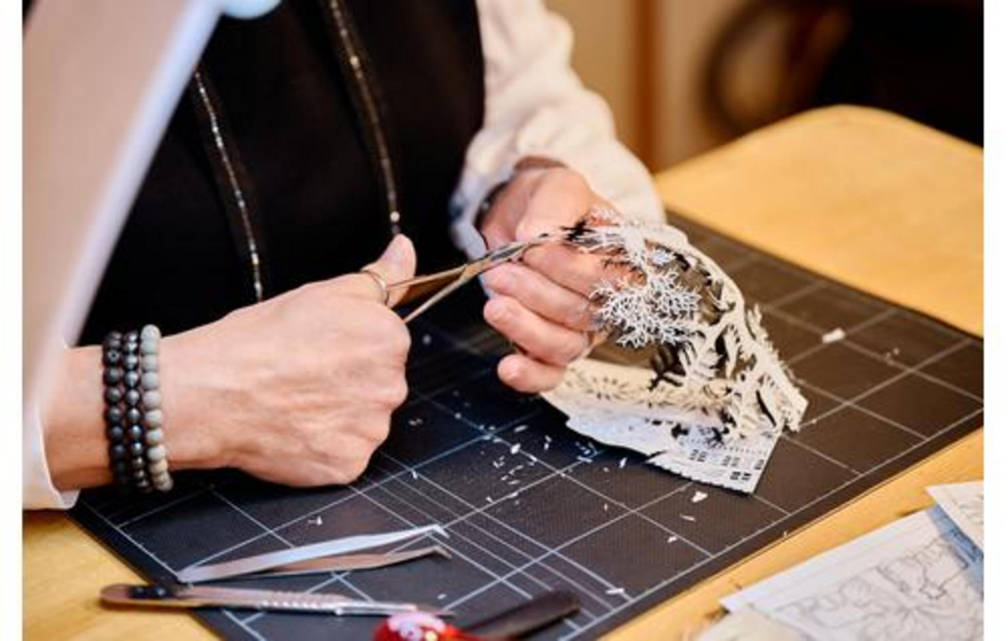 Mani che tagliano intricata arte di carta bianca con forbici su tappetino nero.