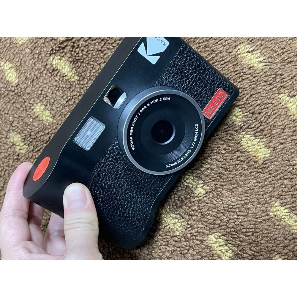 Eine schwarze Kodak Mini Shot 2 Ära Kamera wird auf einem braunen Teppich gehalten.