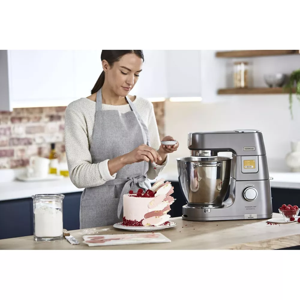 Kobieta dekoruje ciasto jagodami i posypką obok miksera stojącego.