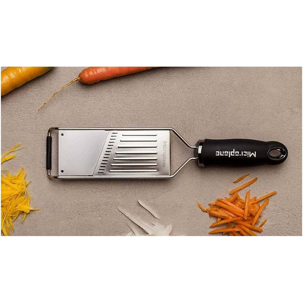Ein Microplane-Hobel mit schwarzem Griff liegt auf grauem Untergrund. Daneben liegen Karottenstreifen.