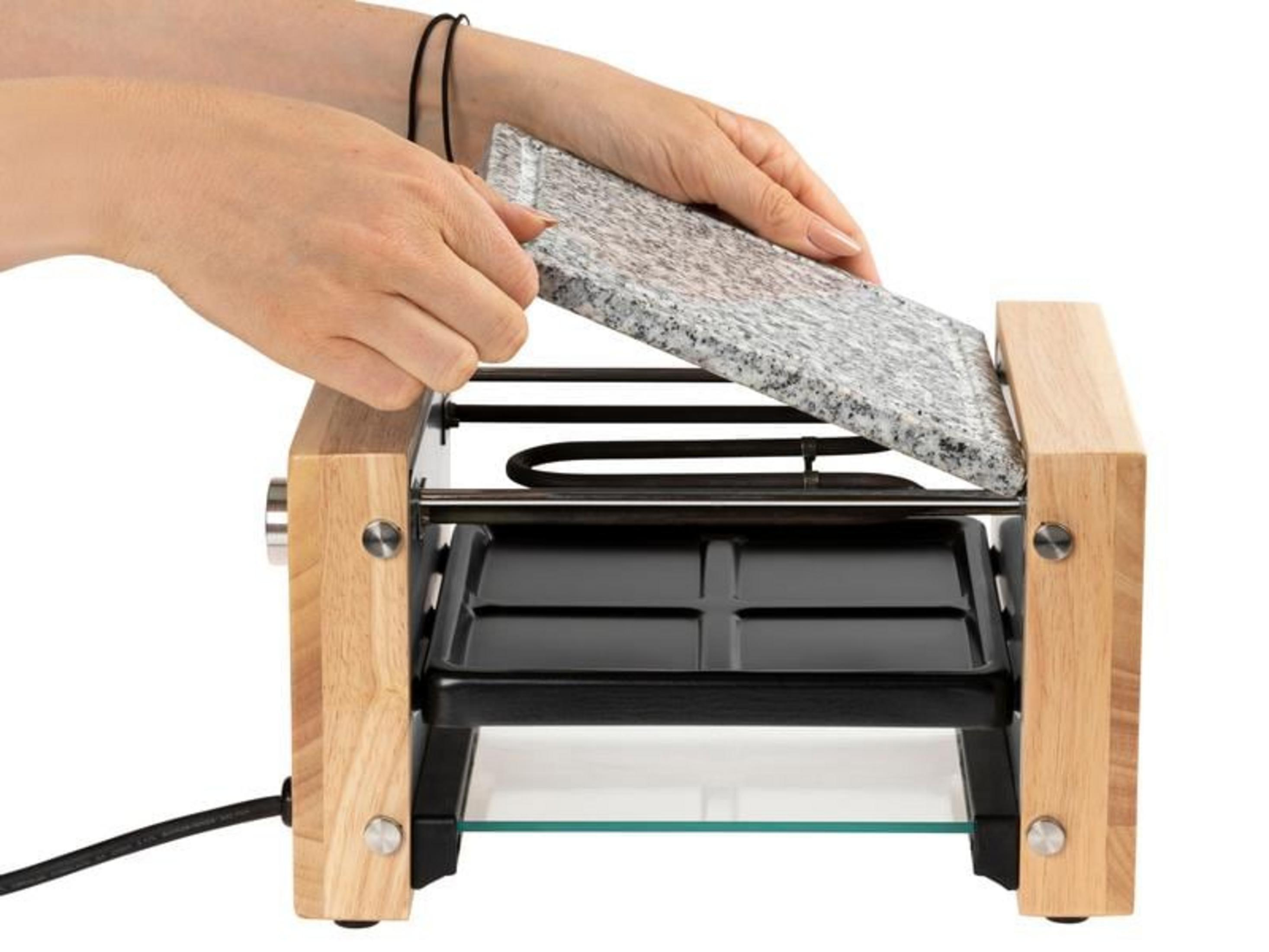 Hände legen Steinplatte auf Raclette-Grill mit Holzrahmen.