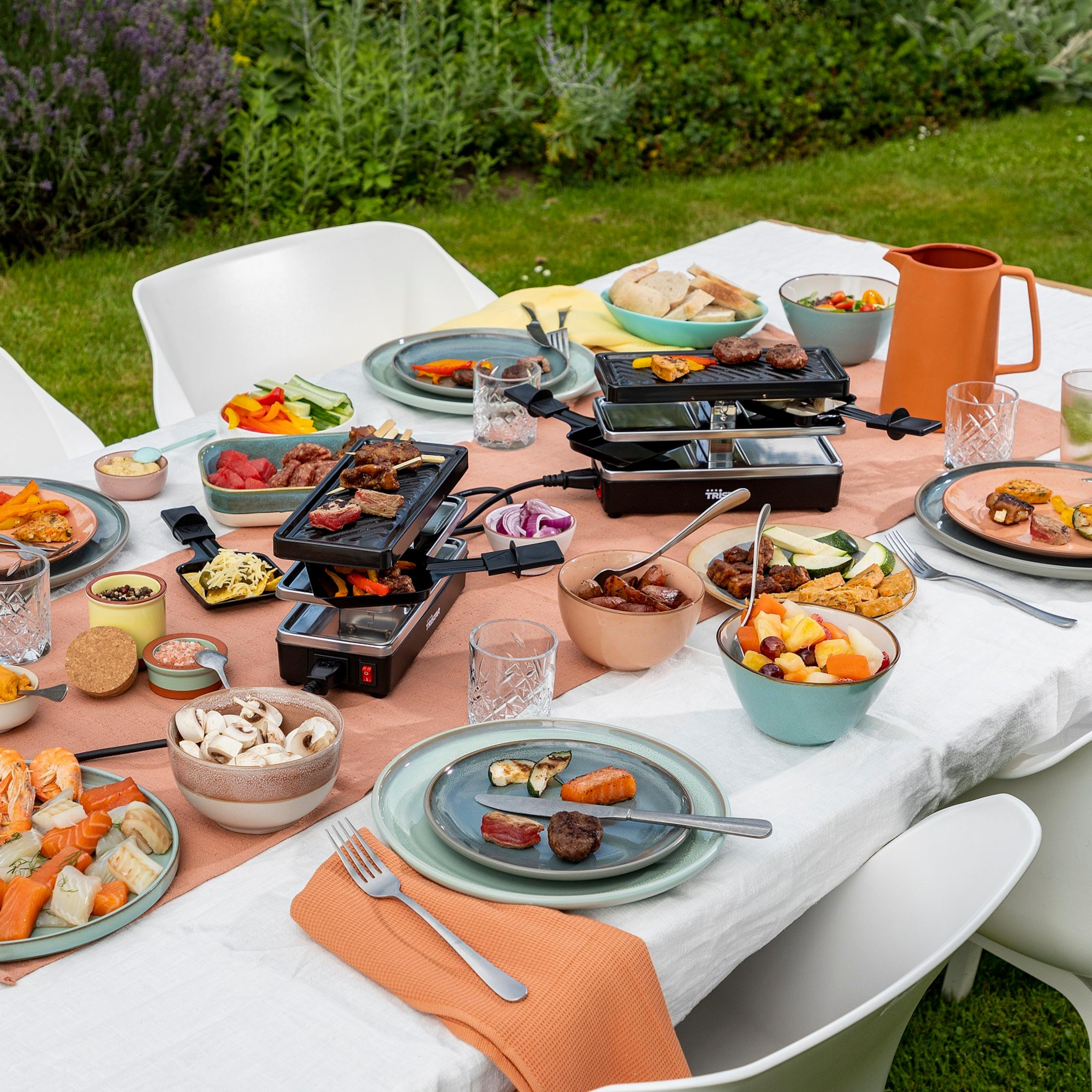 Un tavolo da giardino è apparecchiato con griglie per raclette, carne, pesce, verdure, frutta e pane.
