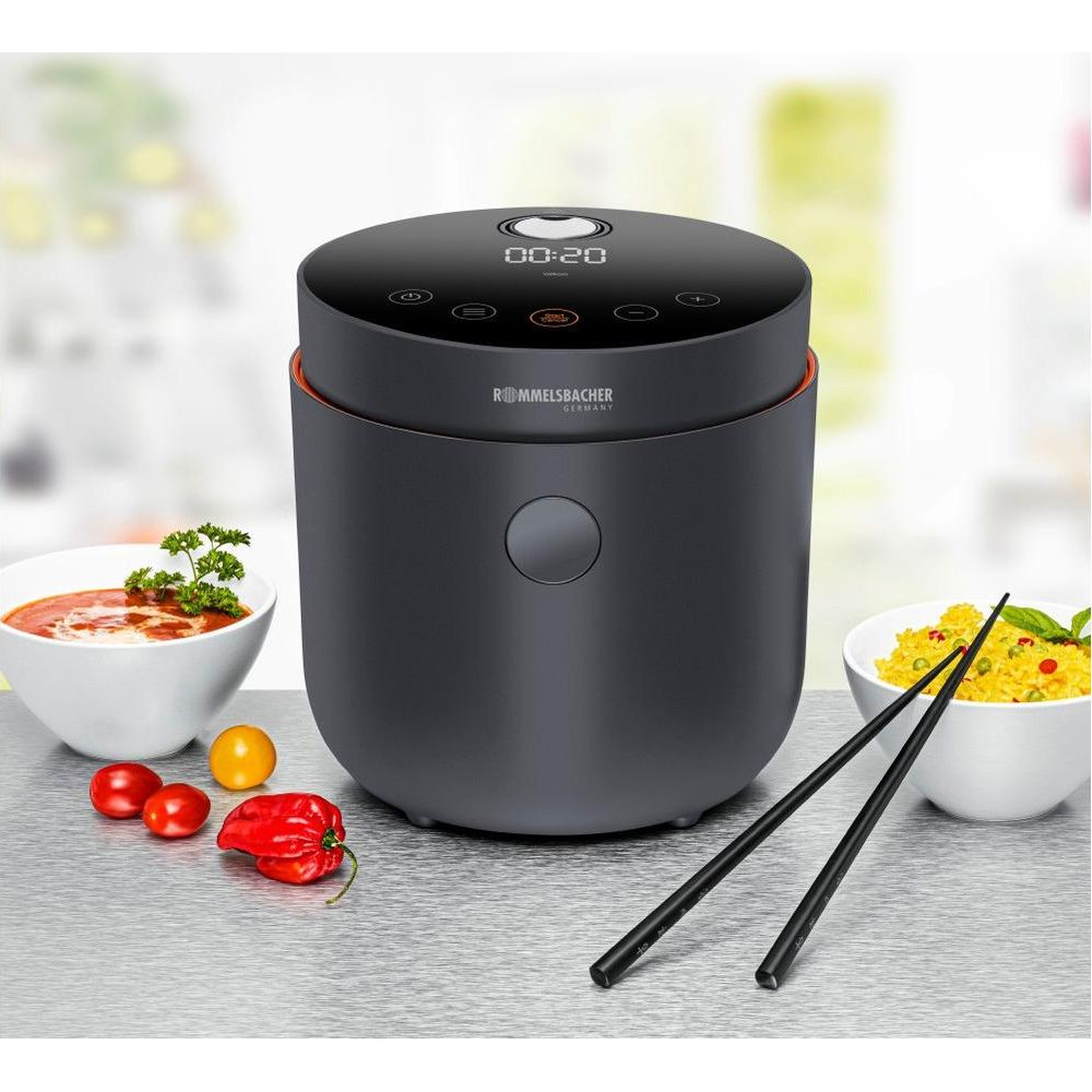 Ciemny garnek do ryżu z czerwonymi akcentami stoi na stole. Obok znajdują się miski z jedzeniem i pałeczki.