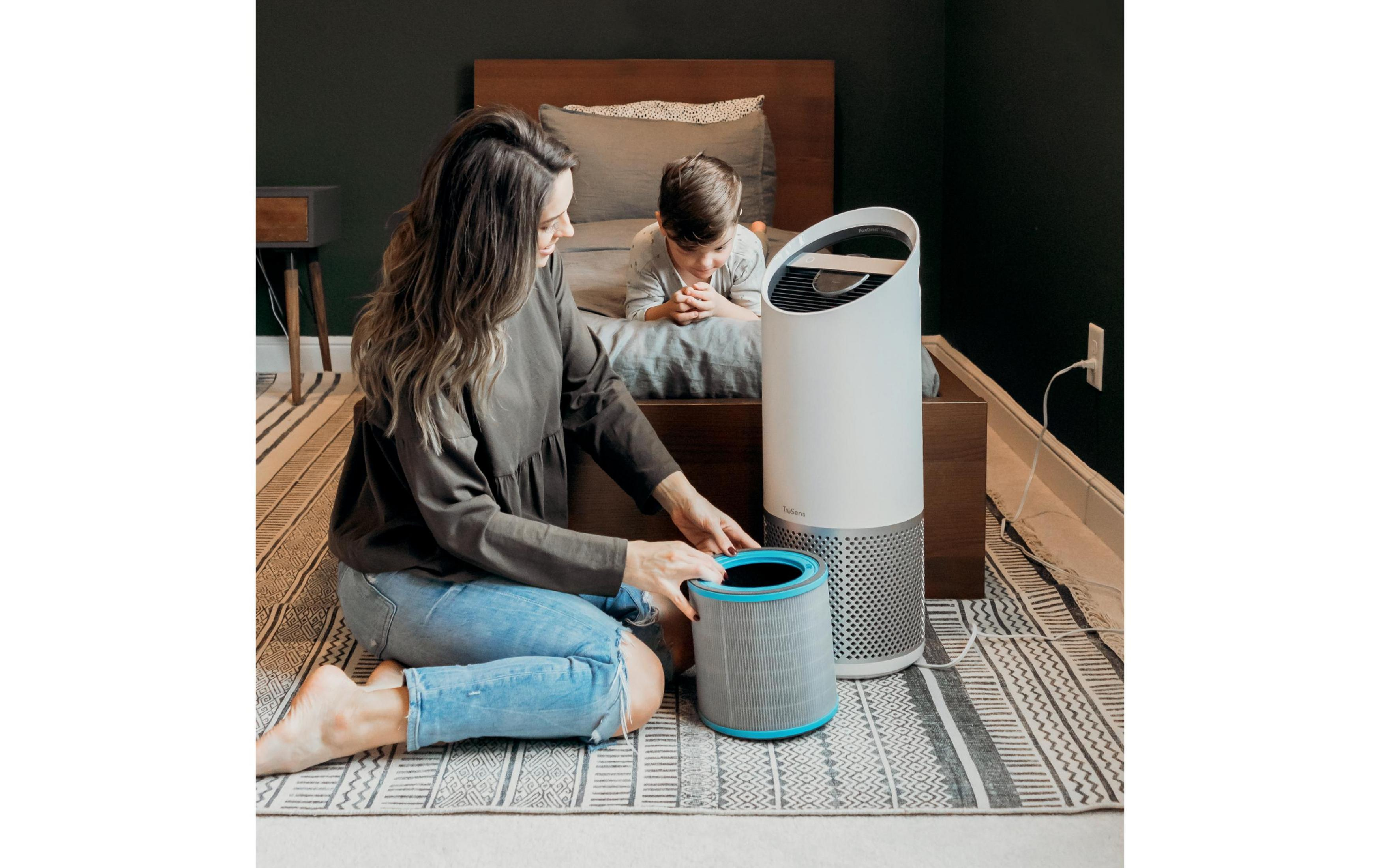 Kobieta wymienia filtr oczyszczacza powietrza obok dziecka na łóżku.