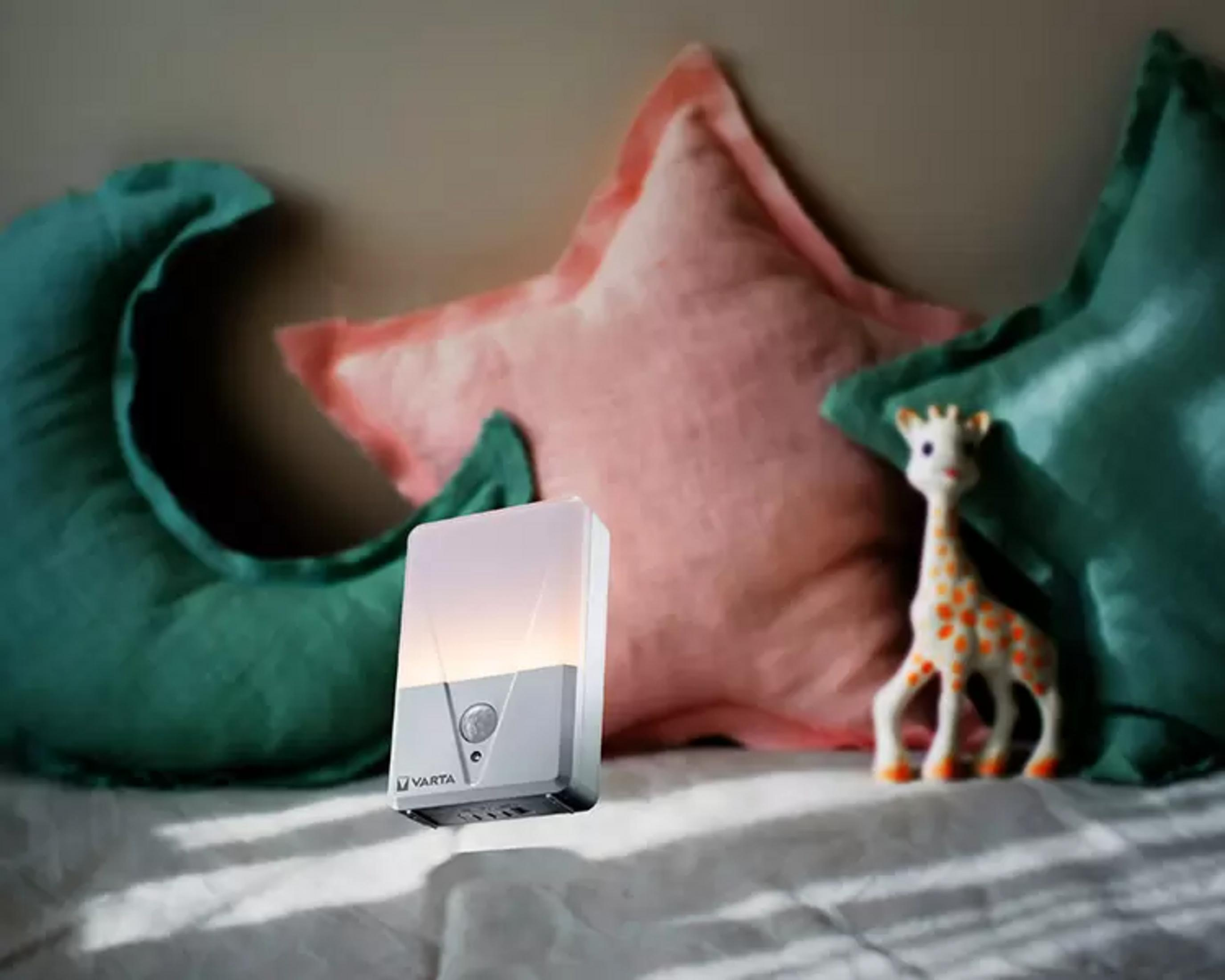 Ein Varta Nachtlicht steht auf einem Bett neben Kissen und einer Giraffe.