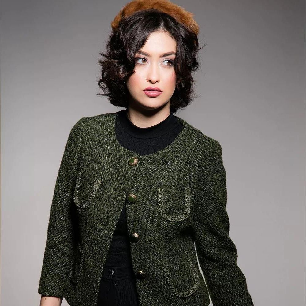 Une femme aux cheveux courts et foncés porte une veste verte en tweed à boutons dorés.