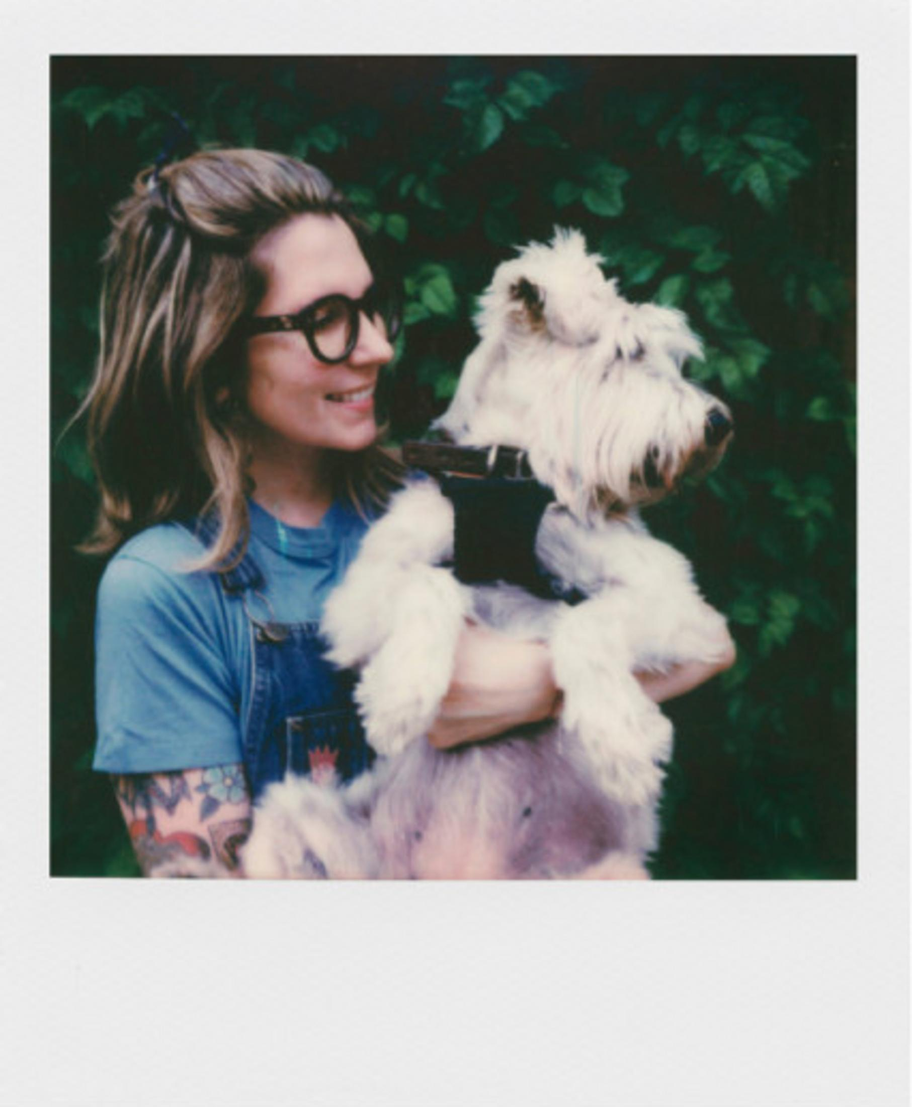 Kobieta w okularach trzyma białego psa na tle ciemnych liści.