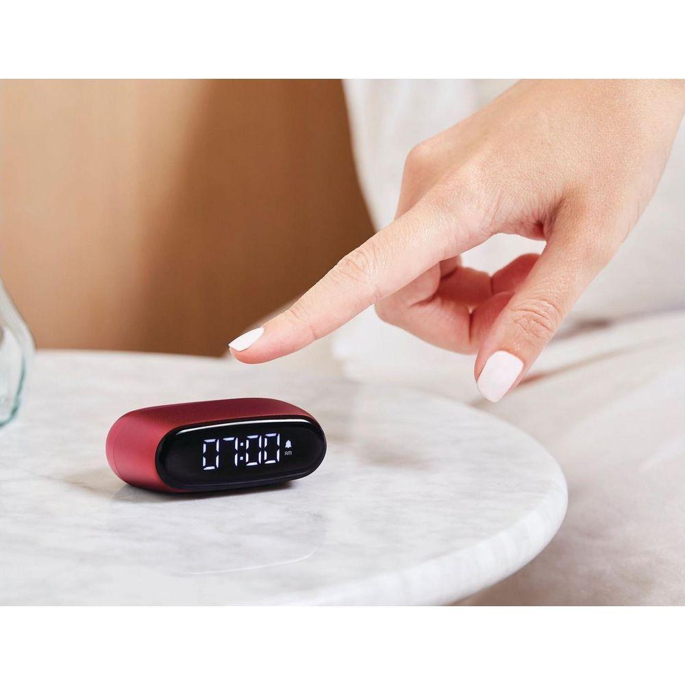 Eine Person berührt einen roten Digitalwecker, der 07:00 Uhr anzeigt.