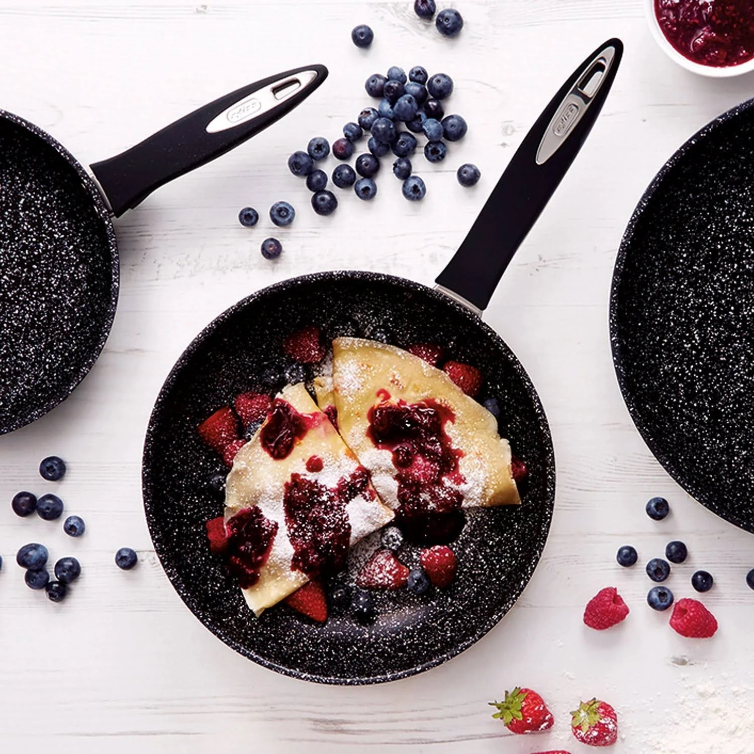 Naleśniki z jagodami i cukrem pudrem na patelni, obok borówek.