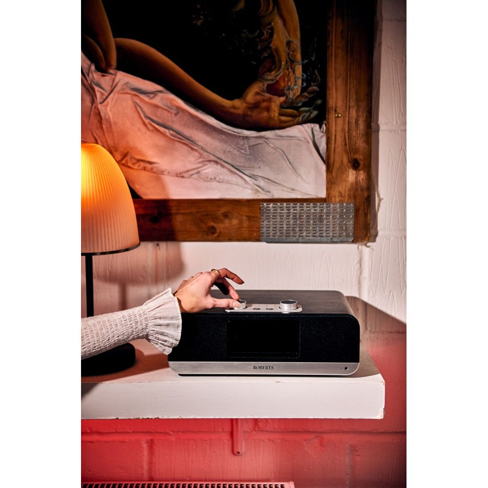 Eine Hand dreht einen Knopf an einem Roberts Radio. Ein Lampenschirm ist links, ein Bild hängt darüber.