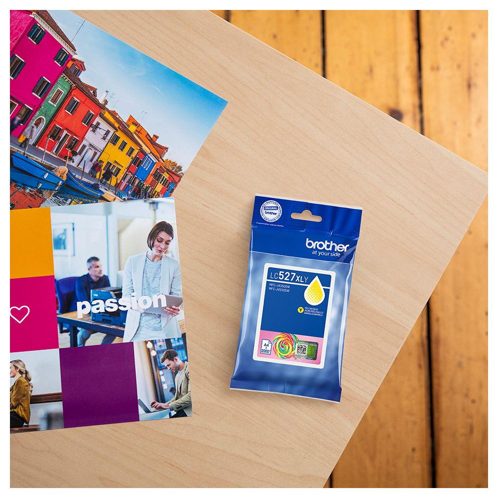 Żółty tusz Brother LC527XLY na drewnie obok wydruków.