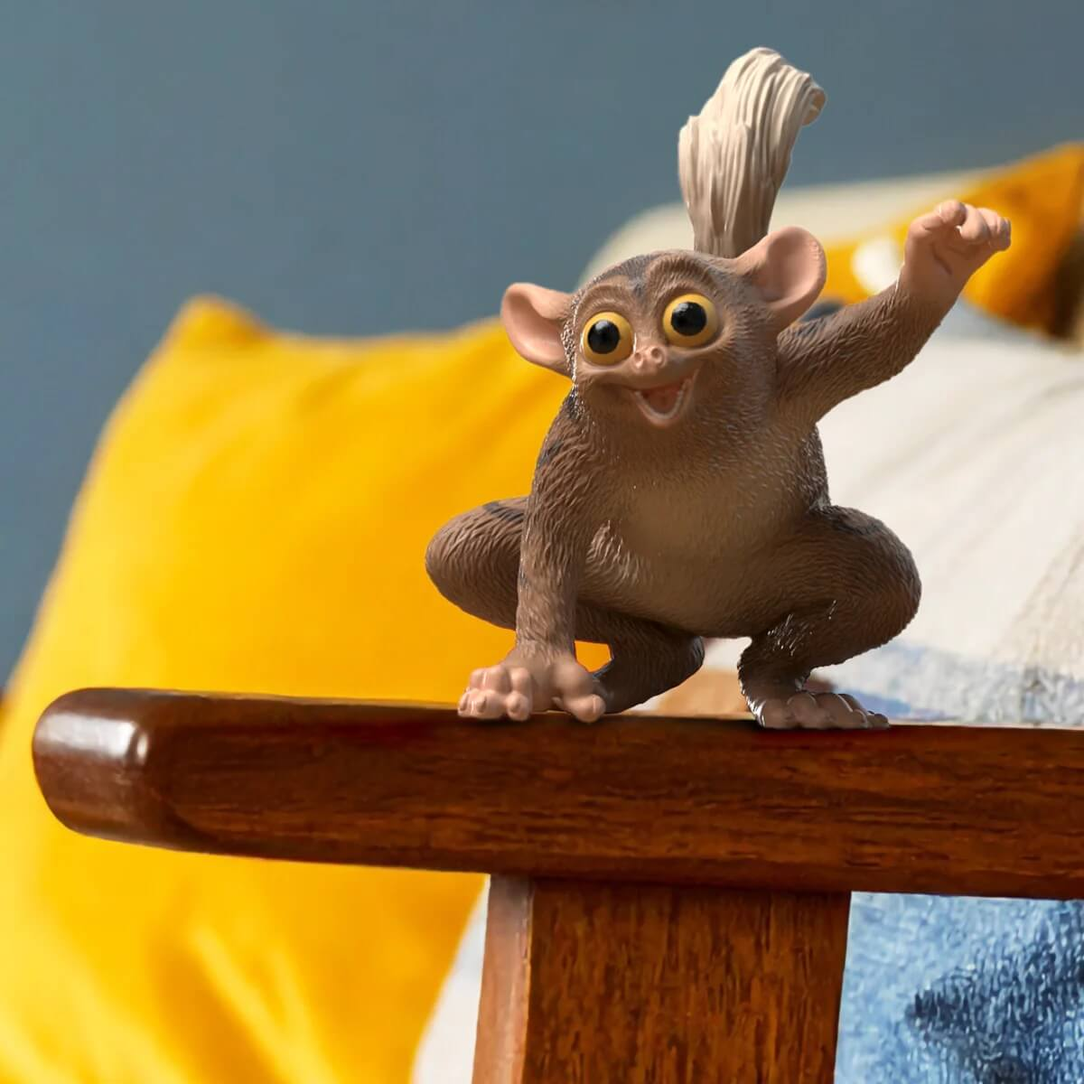 Eine kleine braune Figur eines Koboldmaki sitzt auf einer Holzleiste mit gelben und blauen Kissen.