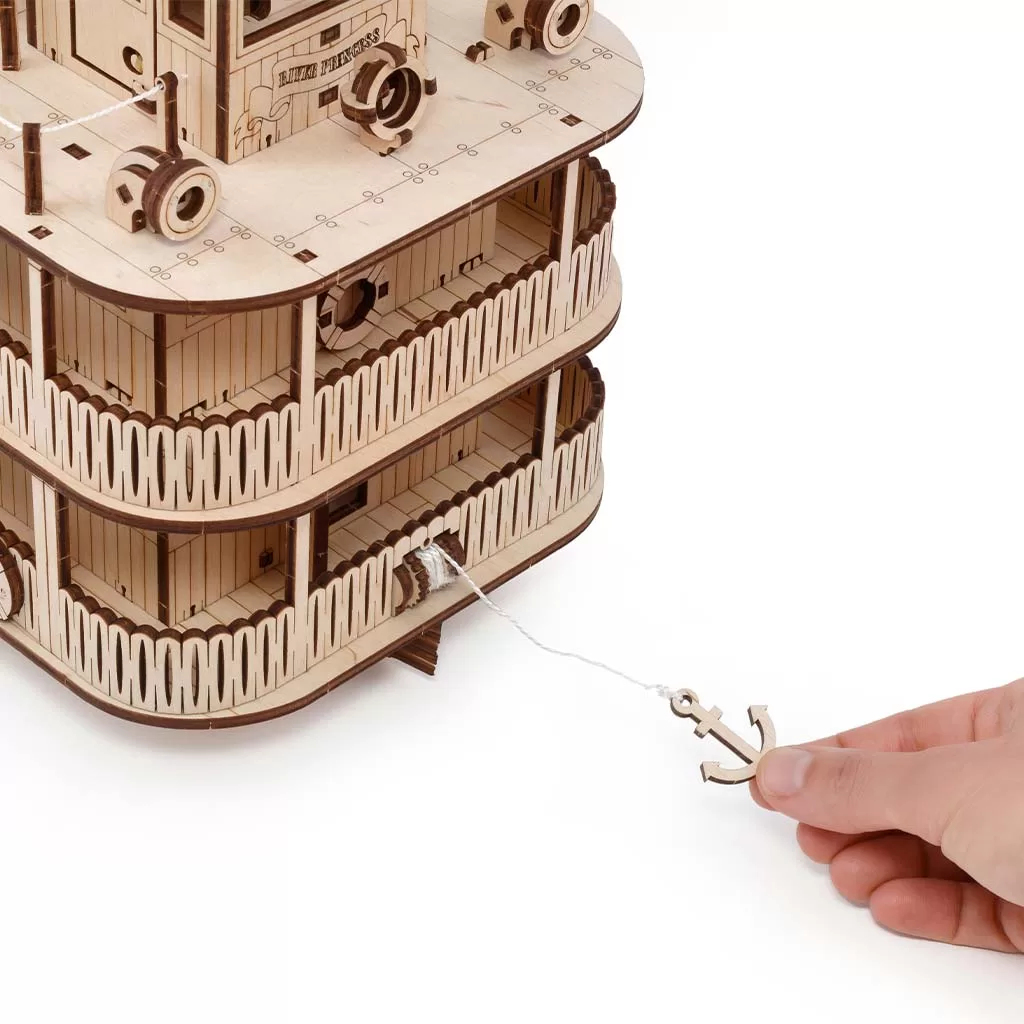 Eine Hand hält einen hölzernen Anker, der an einer Schnur befestigt ist, die von einem Holzmodell eines Dampfschiffes herunterhängt.