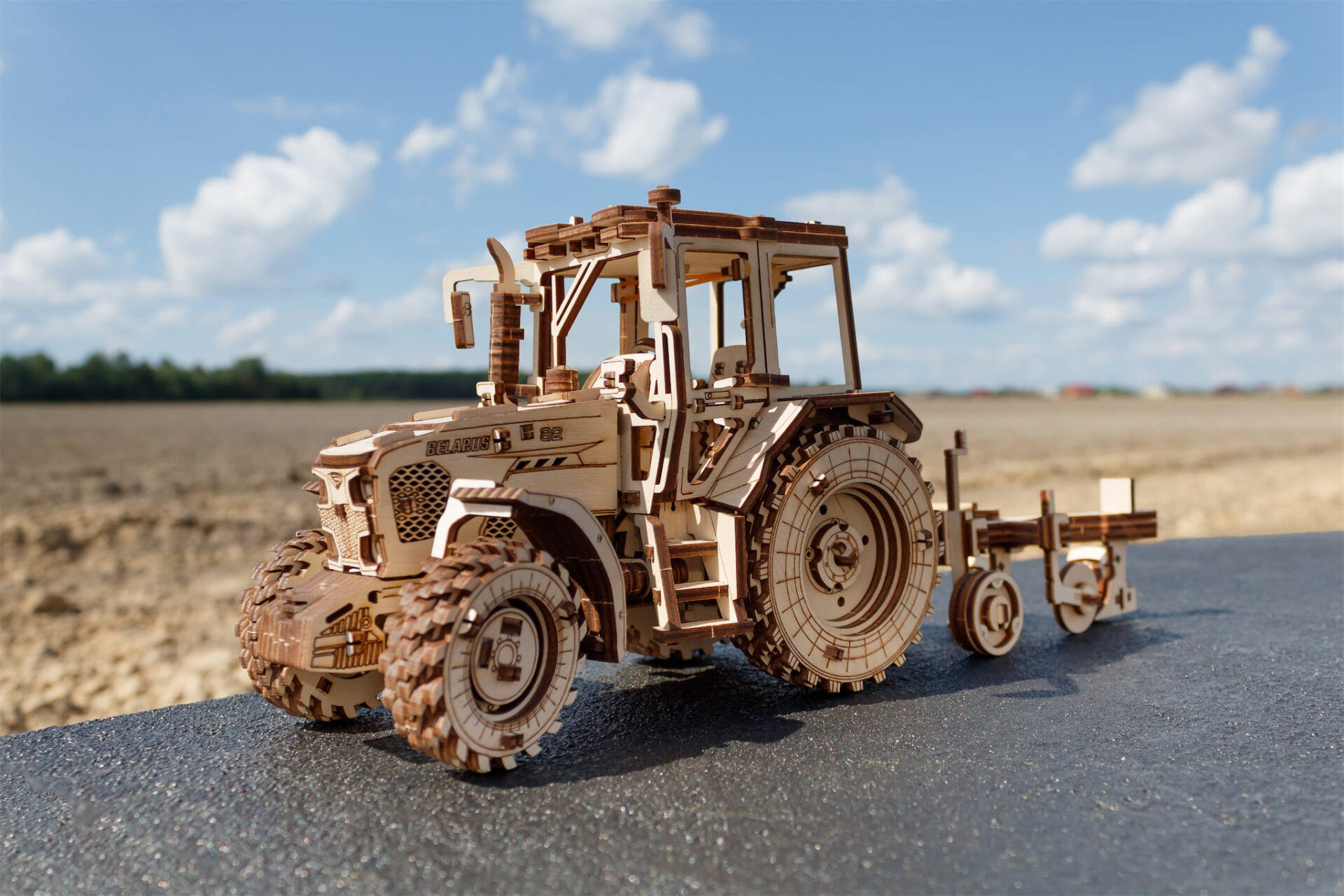 Holzmodell eines Traktors mit Anhänger steht auf Asphalt vor einem Feld.