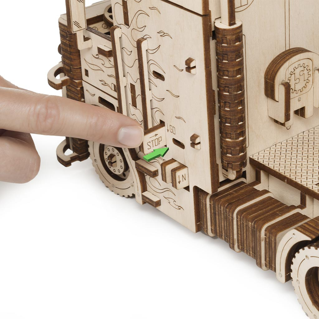 Ein Finger drückt einen Hebel mit 'STOP' auf einem Holzmodell-LKW. Grüne Pfeile zeigen 'STOP' und 'GO'.