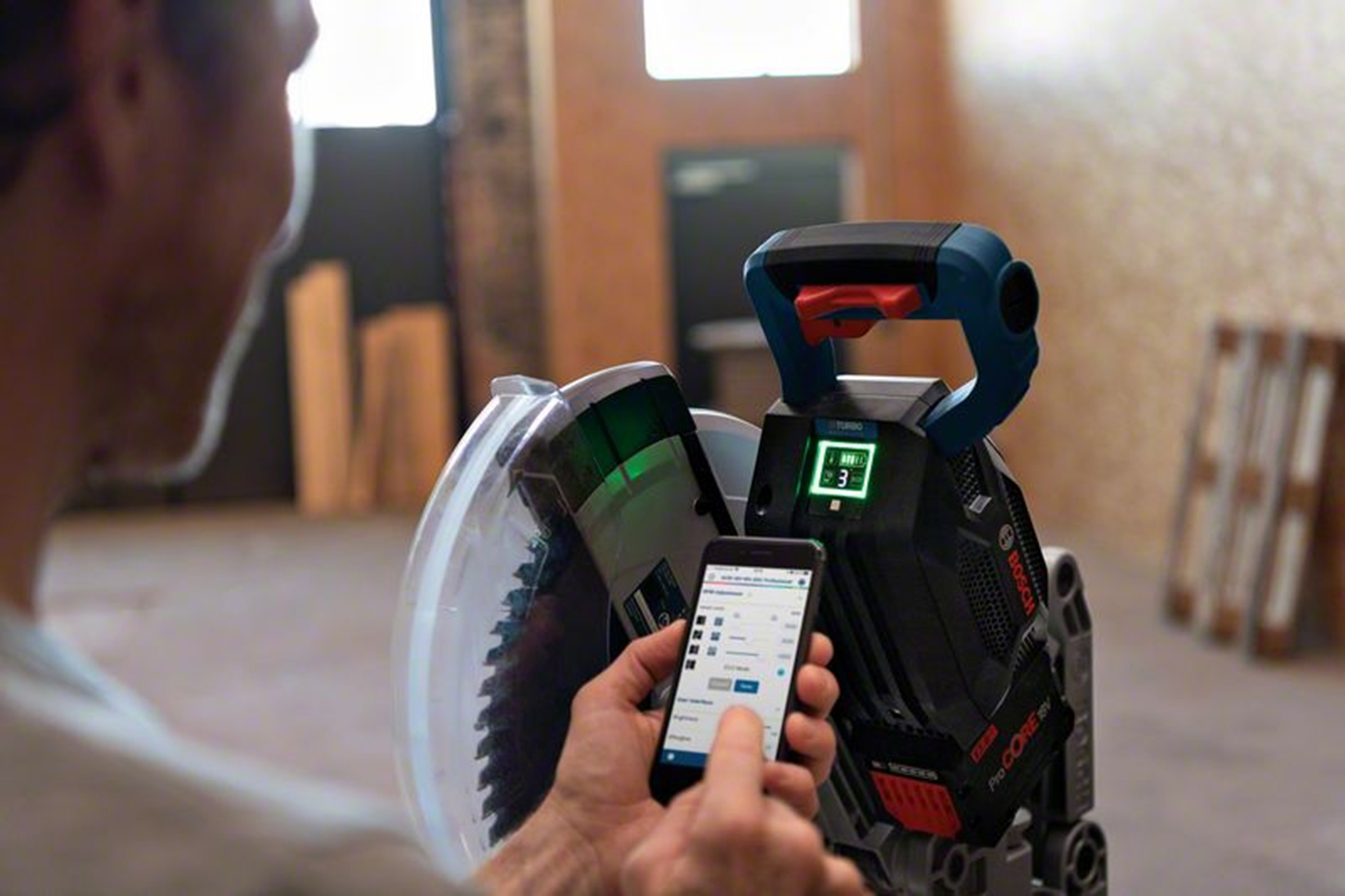 Mann bedient Kreissäge mit Smartphone. Grüne Laserlinie auf Sägeblatt.