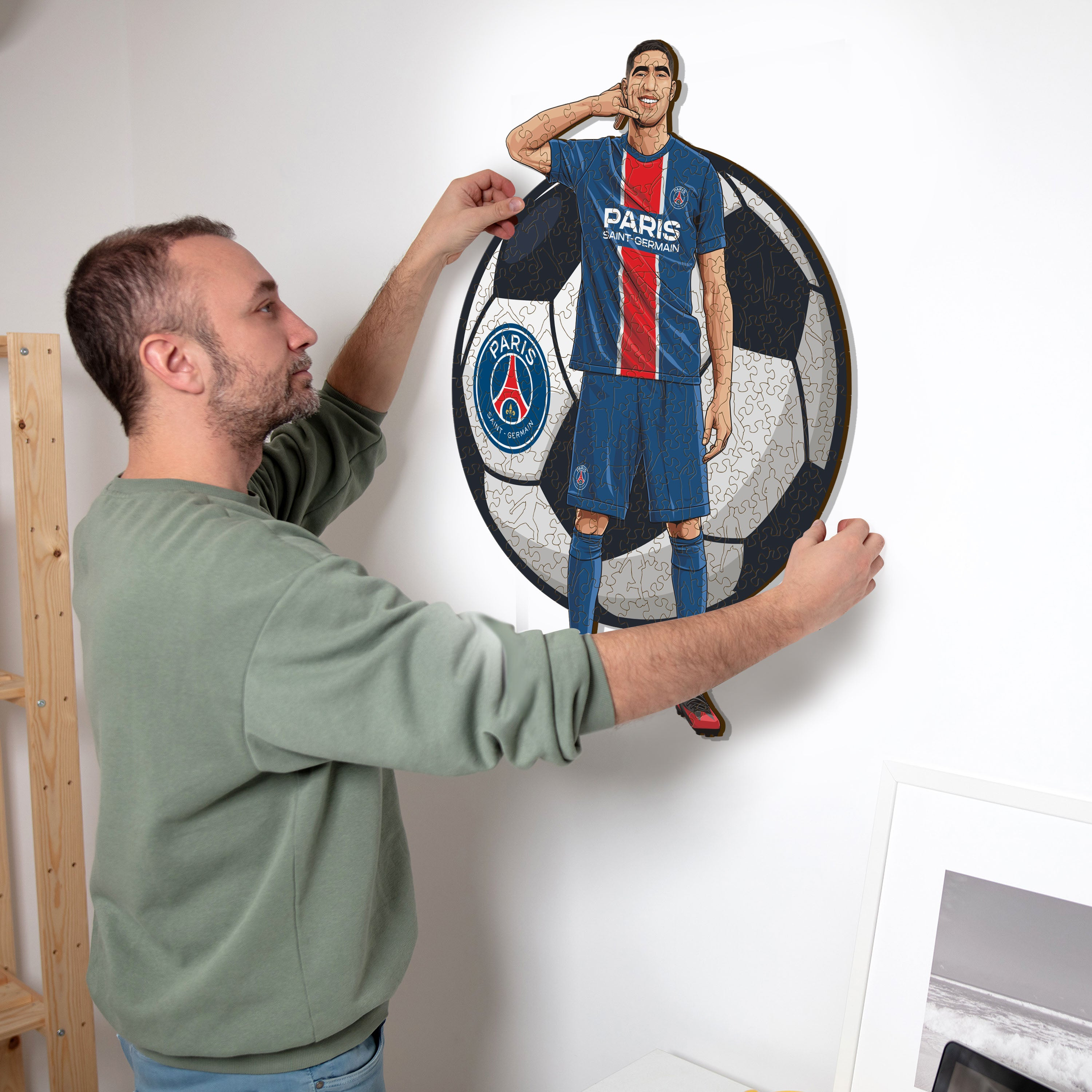 Mann hängt ein Fußball-Puzzle mit einem Spieler in blauer Uniform an die Wand.