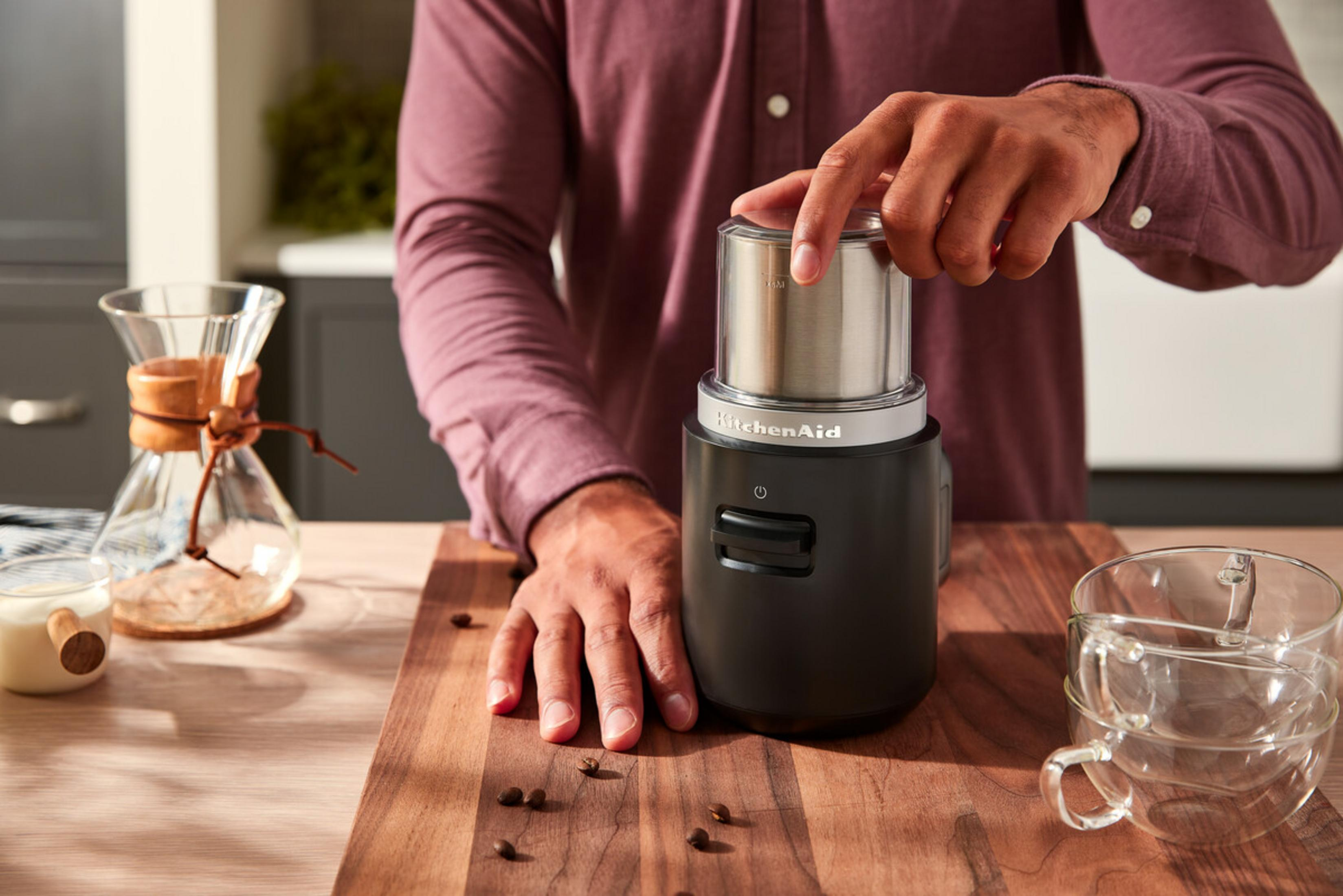 Person mahlt Kaffeebohnen in einer schwarzen KitchenAid-Mühle mit Glasaufsatz.
