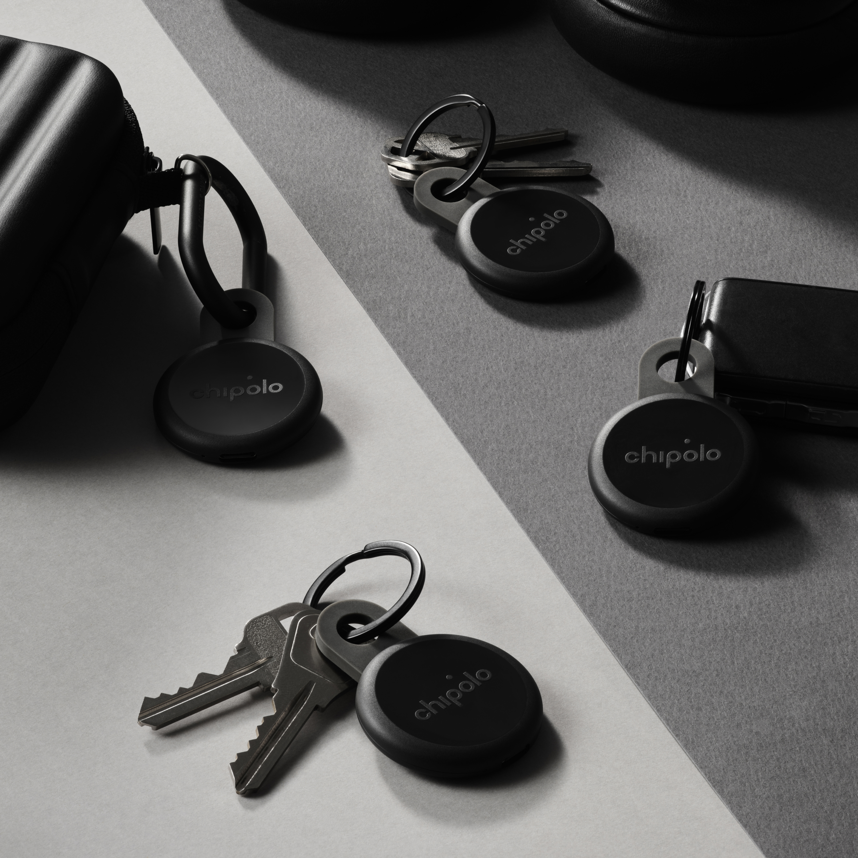 Three black Chipolo keychains with keys on gray and white surfaces.