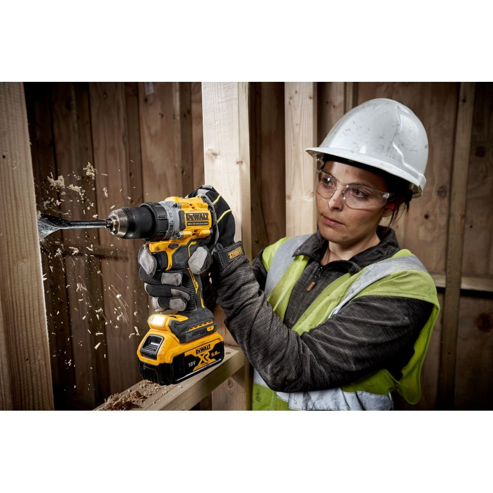 Mujer con casco perfora madera con taladro inalámbrico amarillo. Salta serrín.