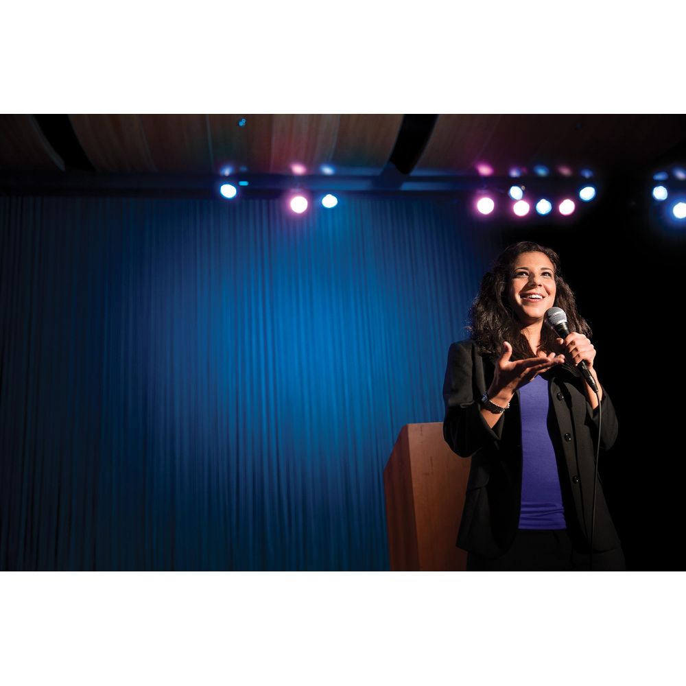 Kobieta mówi do mikrofonu na scenie z niebieskimi zasłonami.