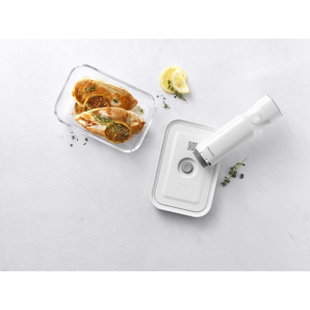 Dos pechugas de pollo con limón y hierbas en un recipiente de vidrio junto a una envasadora al vacío.