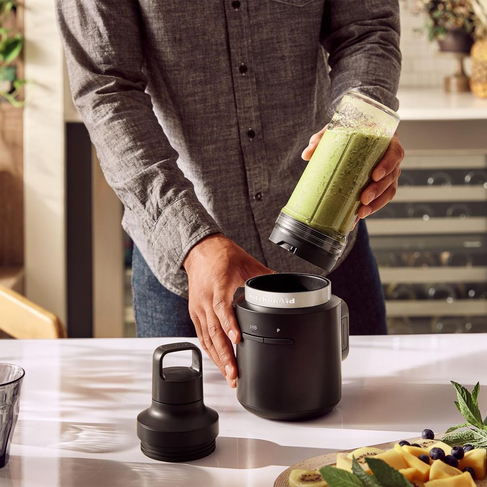 Osoba montuje blender, trzyma kubek z zielonym smoothie nad podstawą. Owoce na stole.